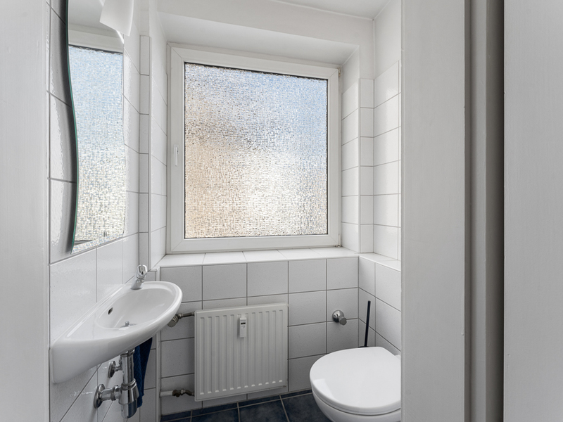 Kleines, weiß gefliestes Badezimmer mit Fenster, Waschbecken und Toilette.