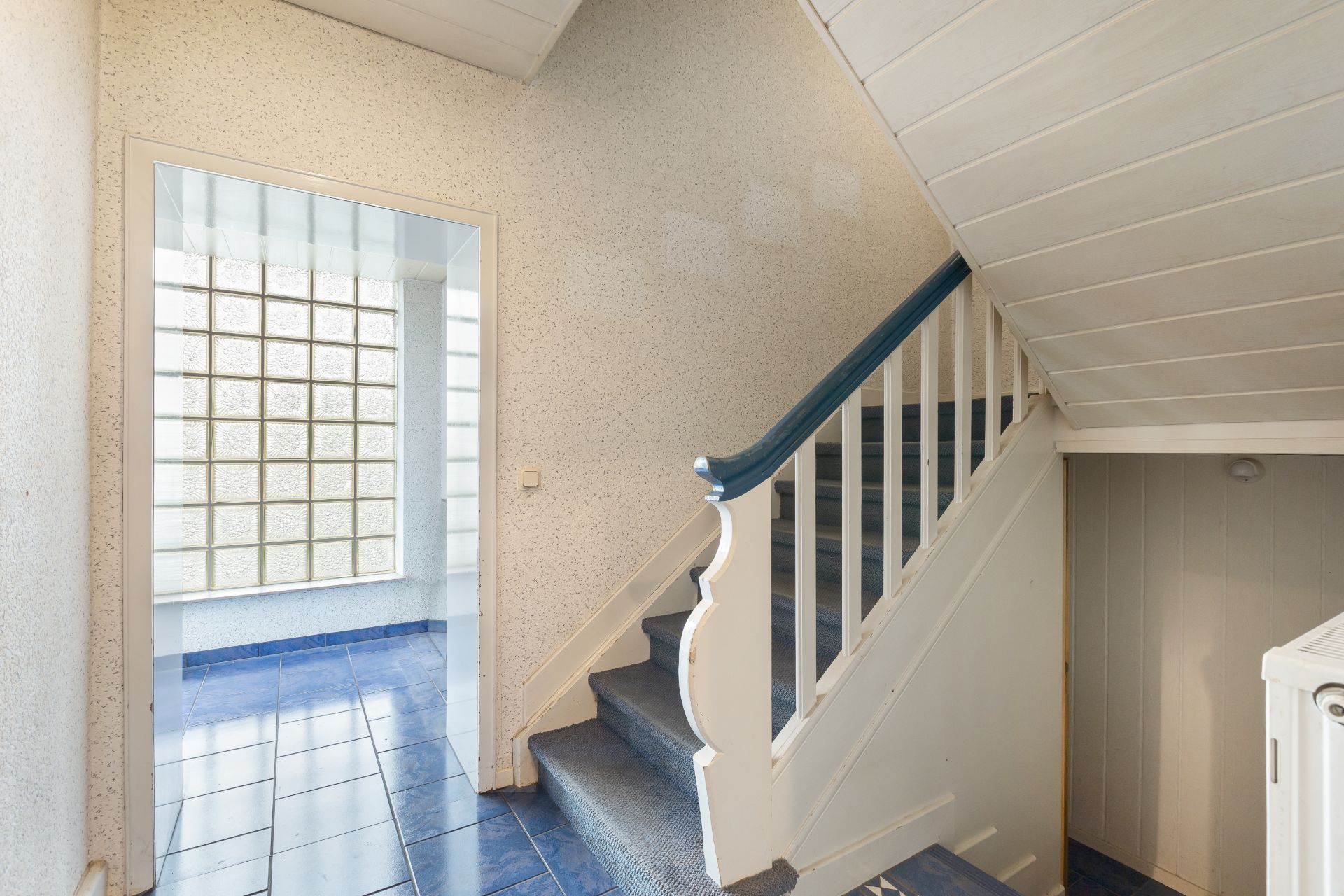 Flur mit blau gefliesten Boden und Treppe, daneben Tür zu einem Raum mit Glasbausteinwand.