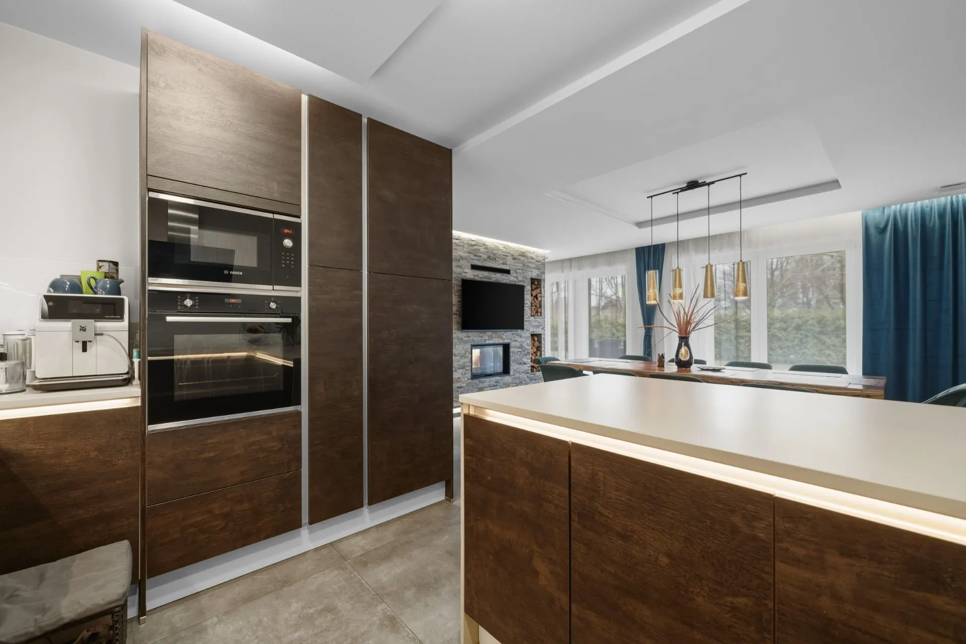 Moderne Küche mit Holzmöbeln, Einbauherd, Kaffeemaschine und Blick ins Wohnzimmer mit Esstisch.