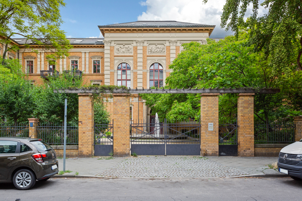 Historisches Wohngebäude mit Ziegelfassade, Tor und begrüntem Vorgarten, davor parkende Autos.