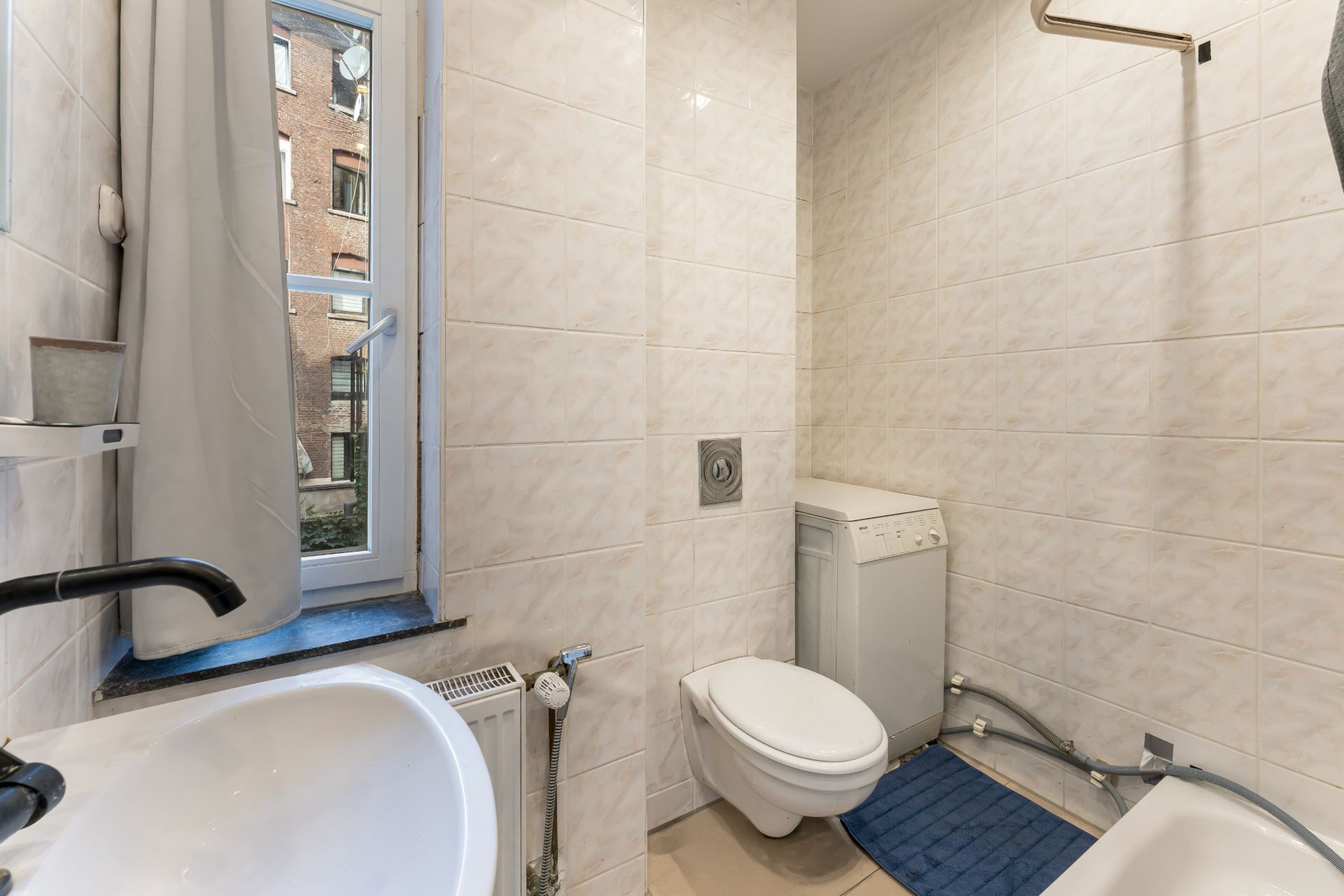 Kleines Badezimmer mit Waschbecken, WC, Waschmaschine, Fenster und Duschbereich.