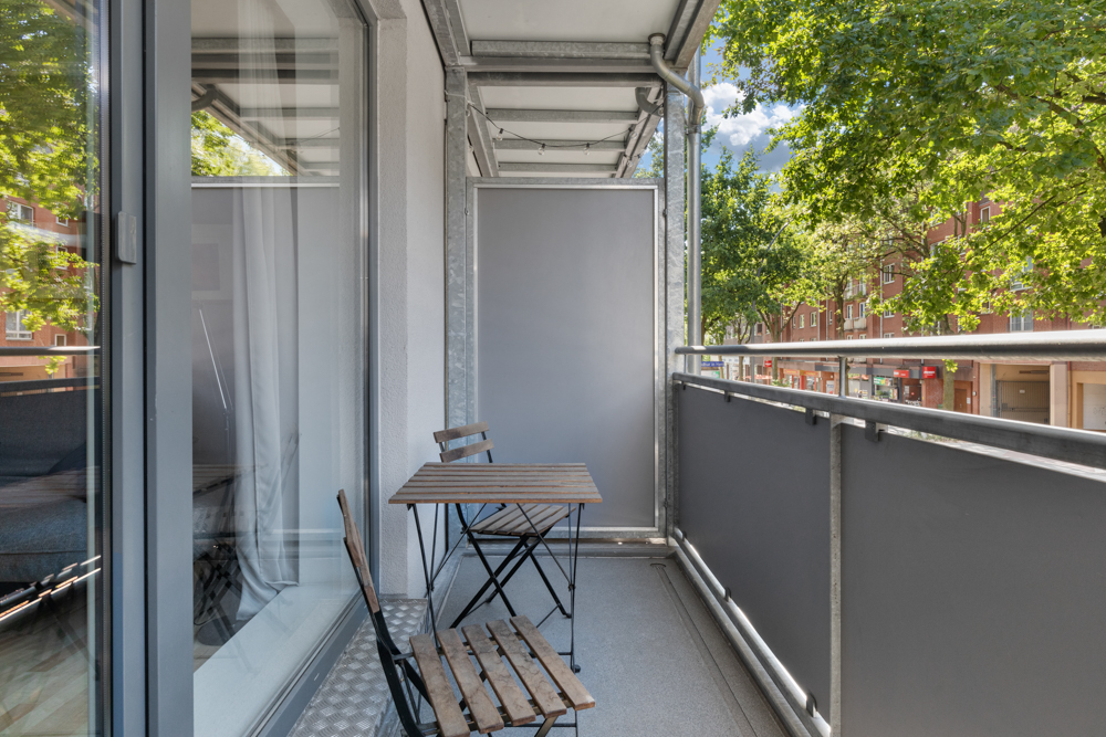 Balkon mit zwei Holzstühlen und einem Tisch, Glasgeländer und Blick auf Bäume.
