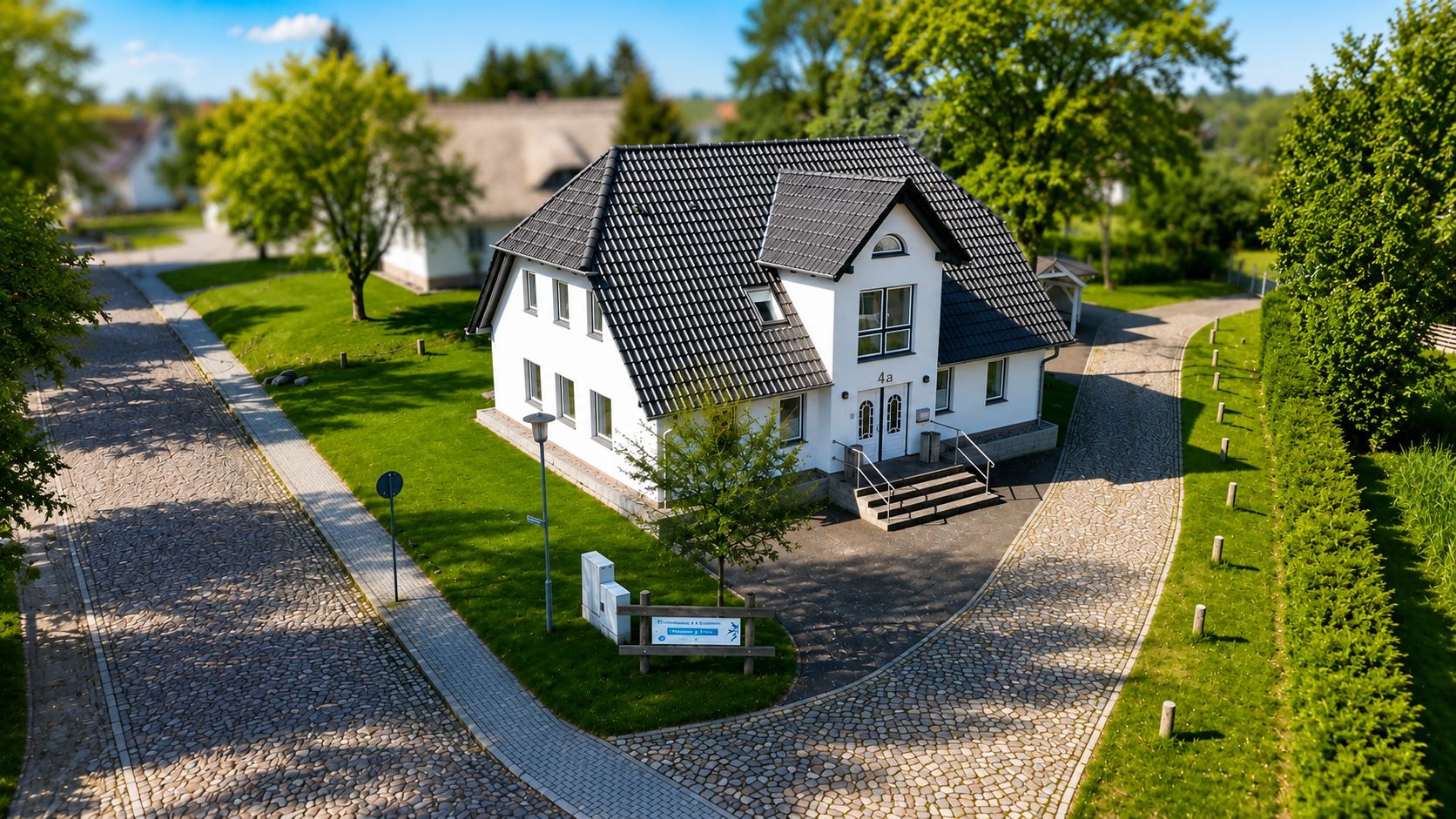 Weißes Einfamilienhaus mit dunklem Dach an einer gepflasterten Straße, umgeben von Bäumen und Grünflächen.