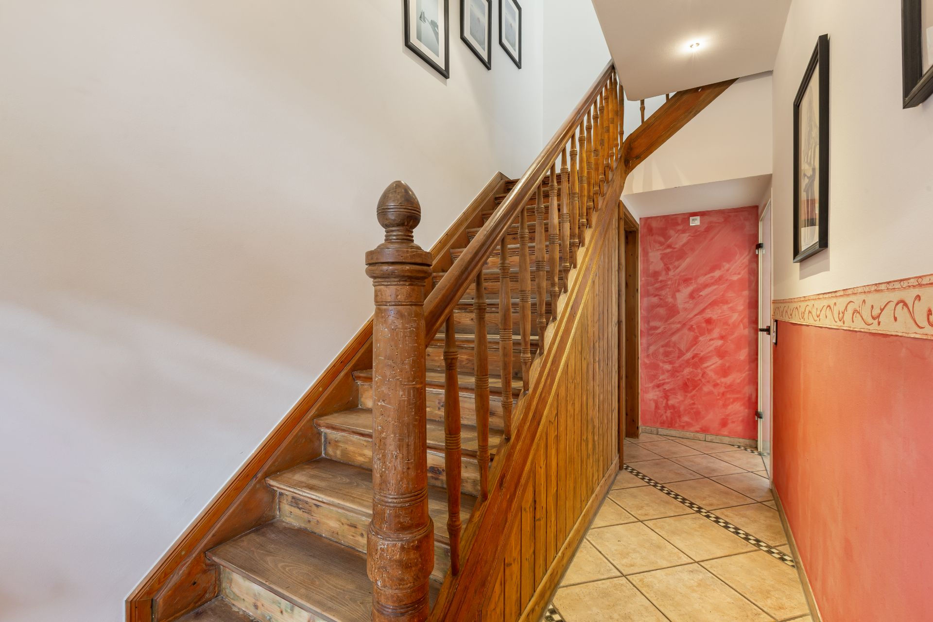 Holztreppe mit Geländer in einem Hausflur mit roten Wänden und Fliesenboden.