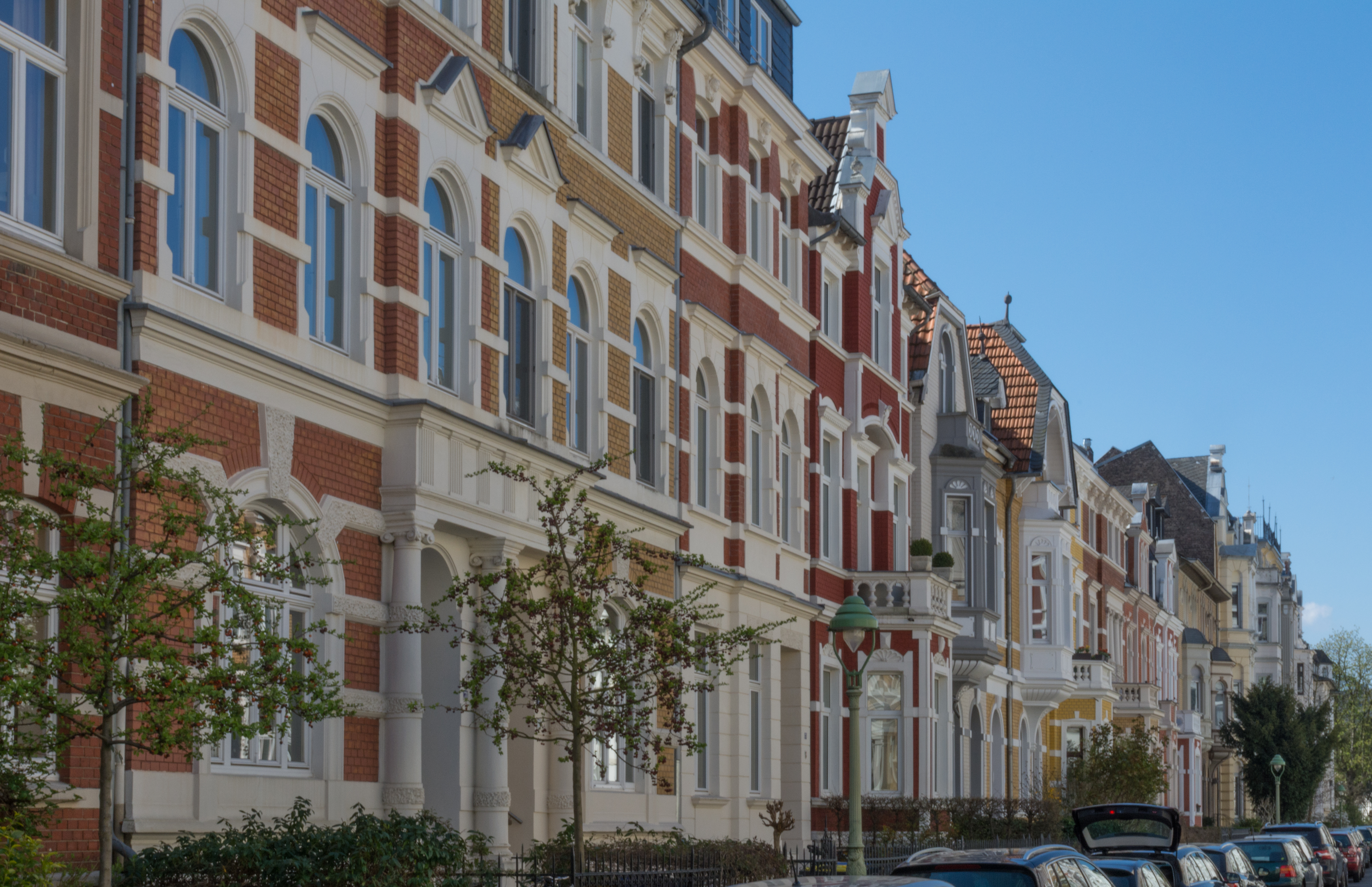 Mehrstöckige Wohnhäuser mit verzierter Fassade und parkenden Autos an einer Straße.