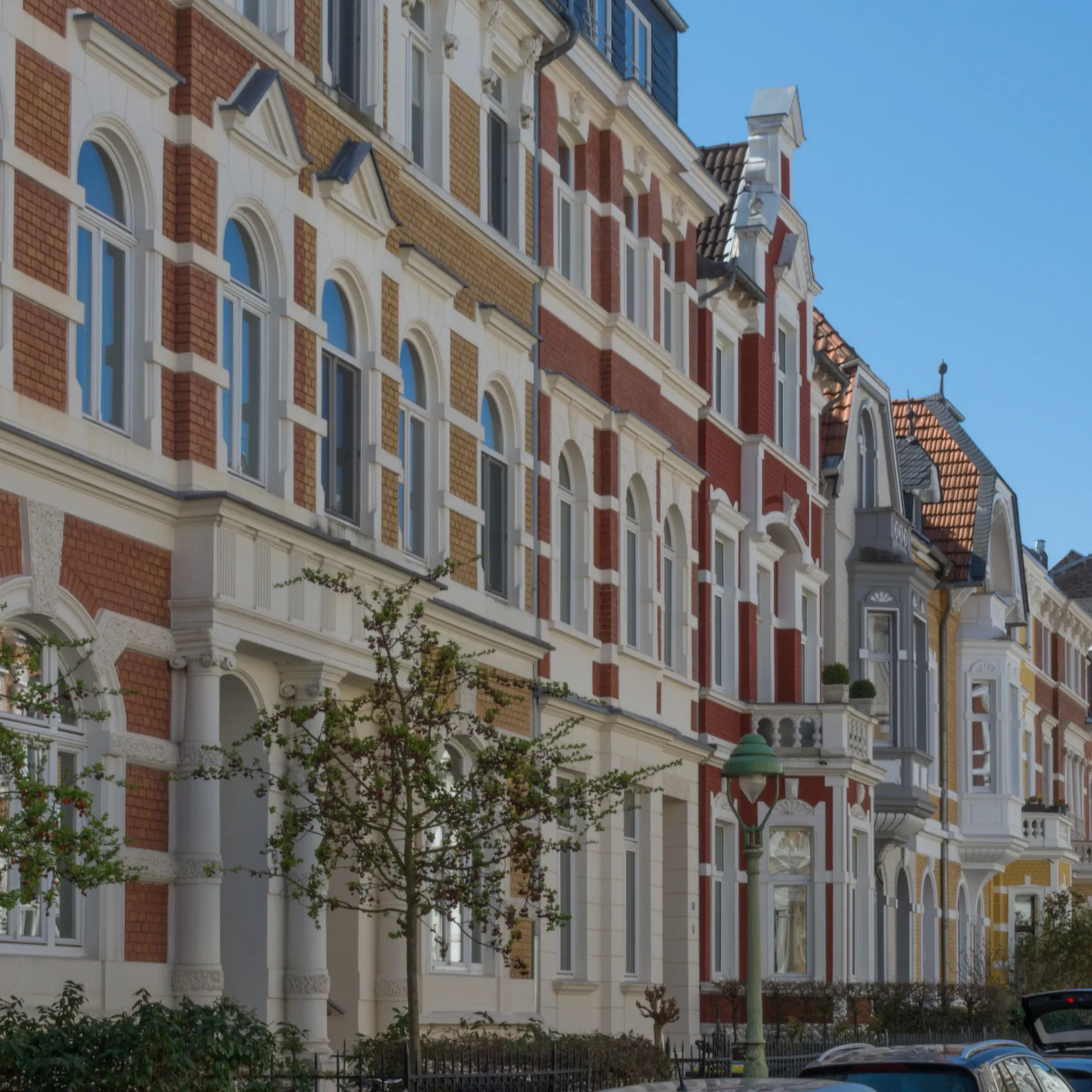 Mehrstöckige Wohnhäuser mit verzierter Fassade und parkenden Autos an einer Straße.