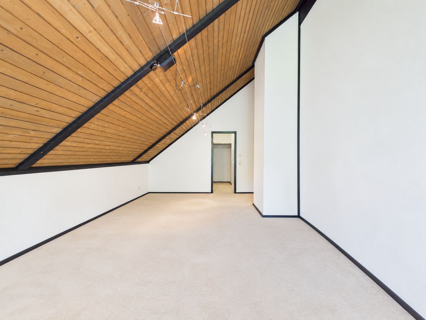 Leerer Dachgeschossraum mit schräger Holzdecke, weißem Teppich und offenem Durchgang.