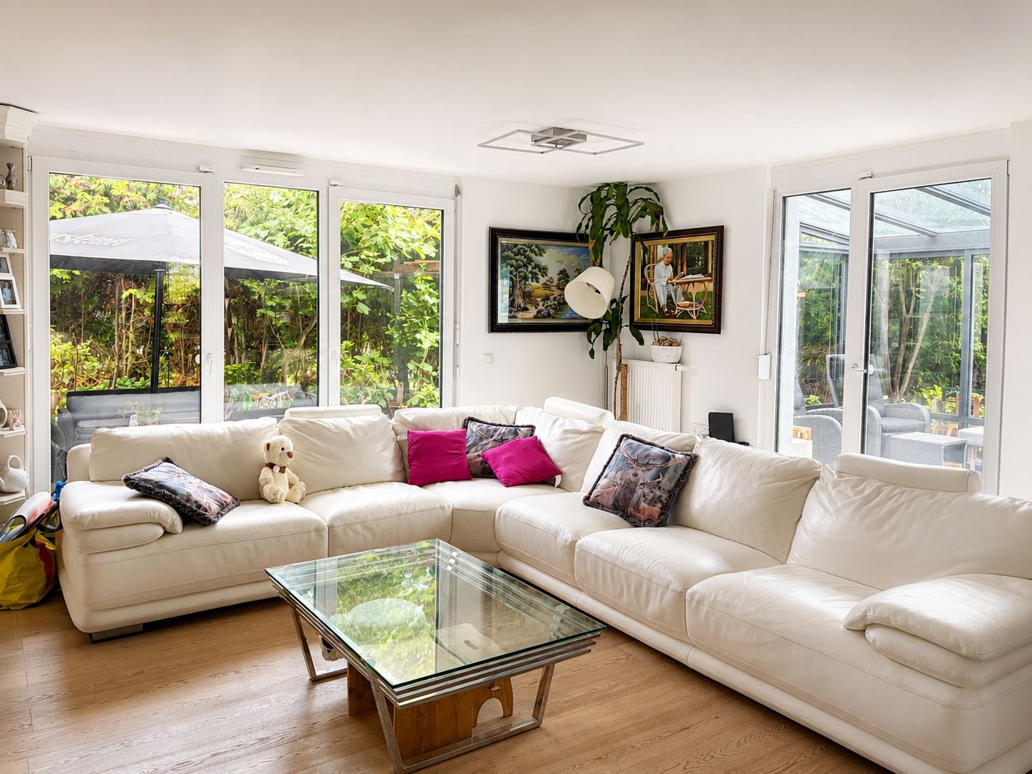 Helles Wohnzimmer mit weißem Ecksofa, Glastisch, Kissen und Fenstern zum Garten.