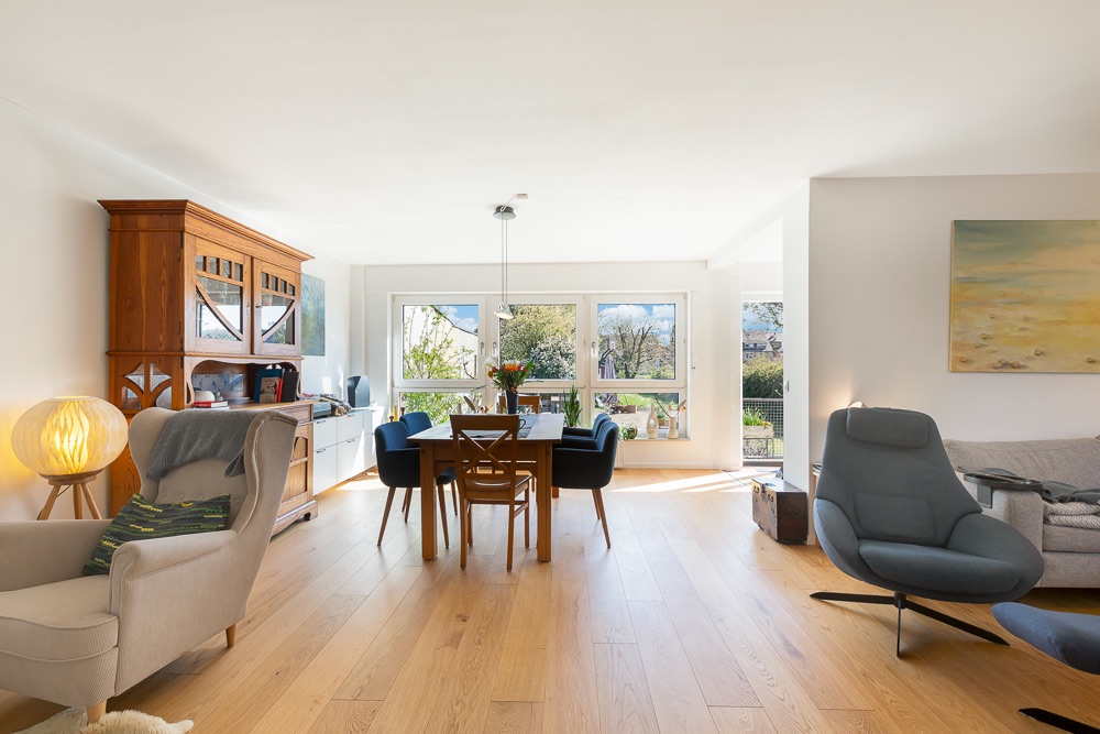 Helles Wohnzimmer mit Holzfußboden, Esstisch, blauen Stühlen und großen Fenstern mit Gartenblick.