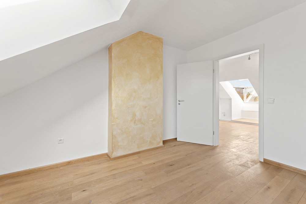 Heller Raum mit Dachschräge, weißer Wand, Holzboden und offener Tür zu weiterem Zimmer.