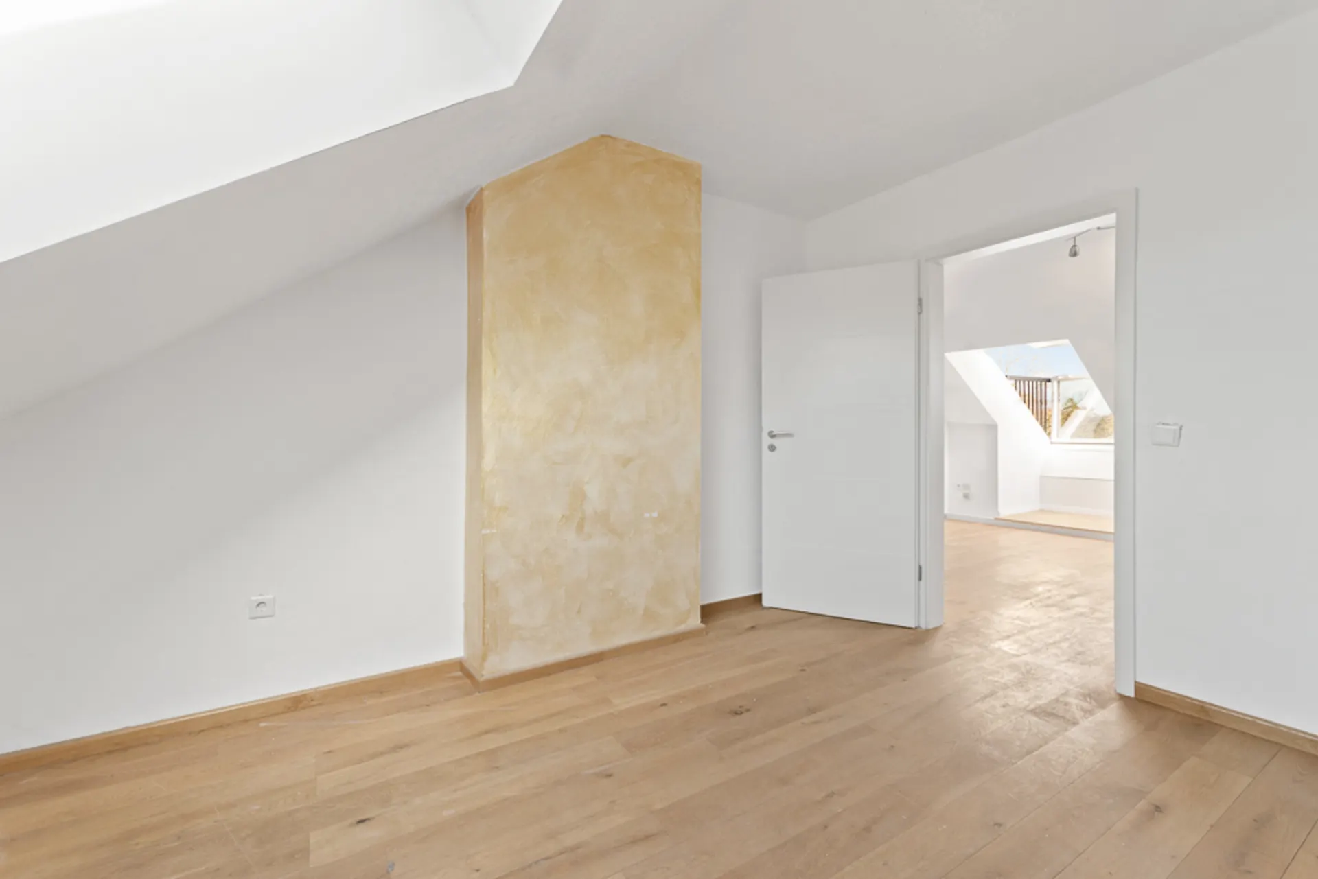 Heller Raum mit Dachschräge, weißer Wand, Holzboden und offener Tür zu weiterem Zimmer.