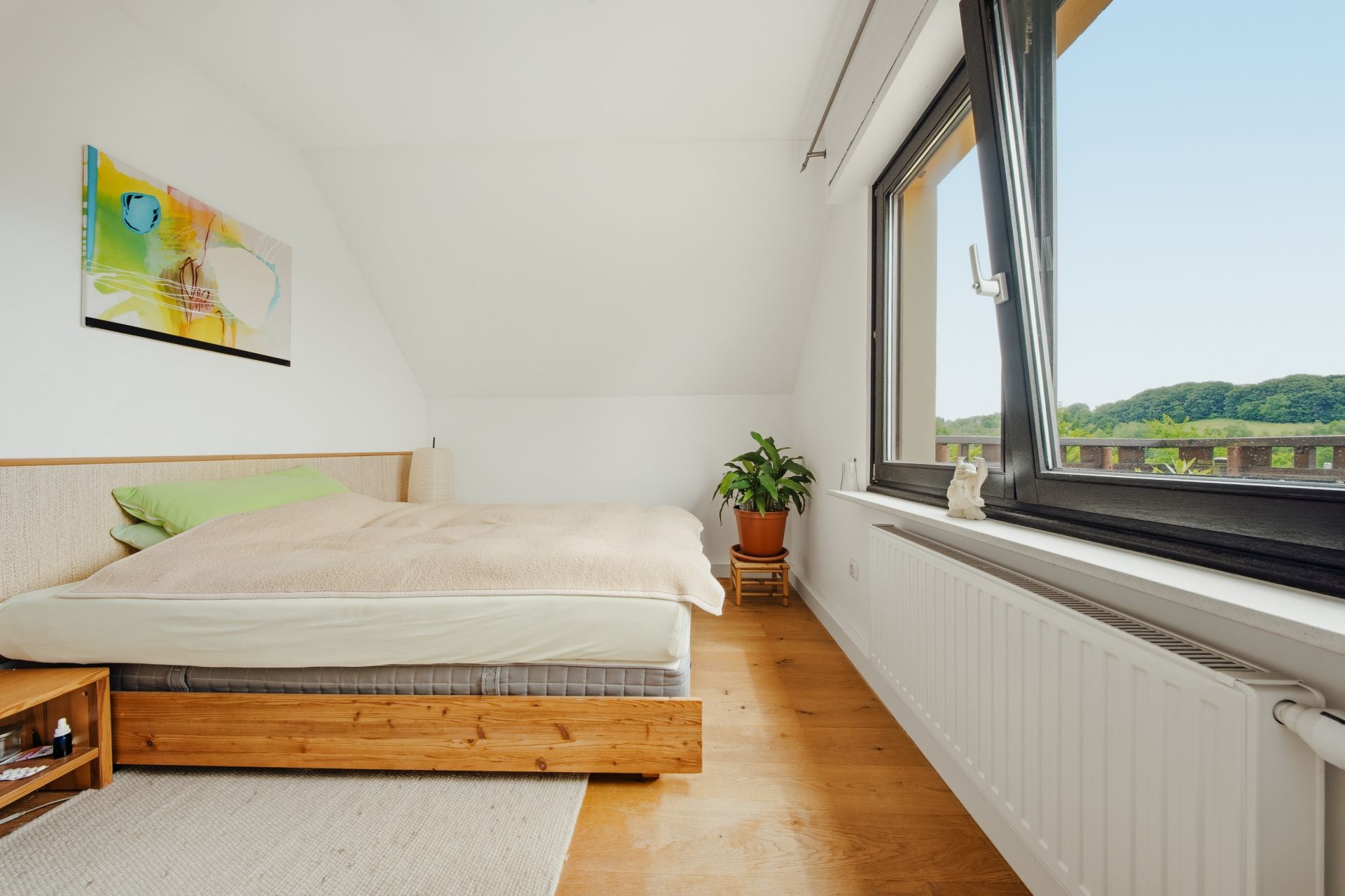 Helles Schlafzimmer mit Holzbett, großem Fenster, Pflanze und Wandbild.