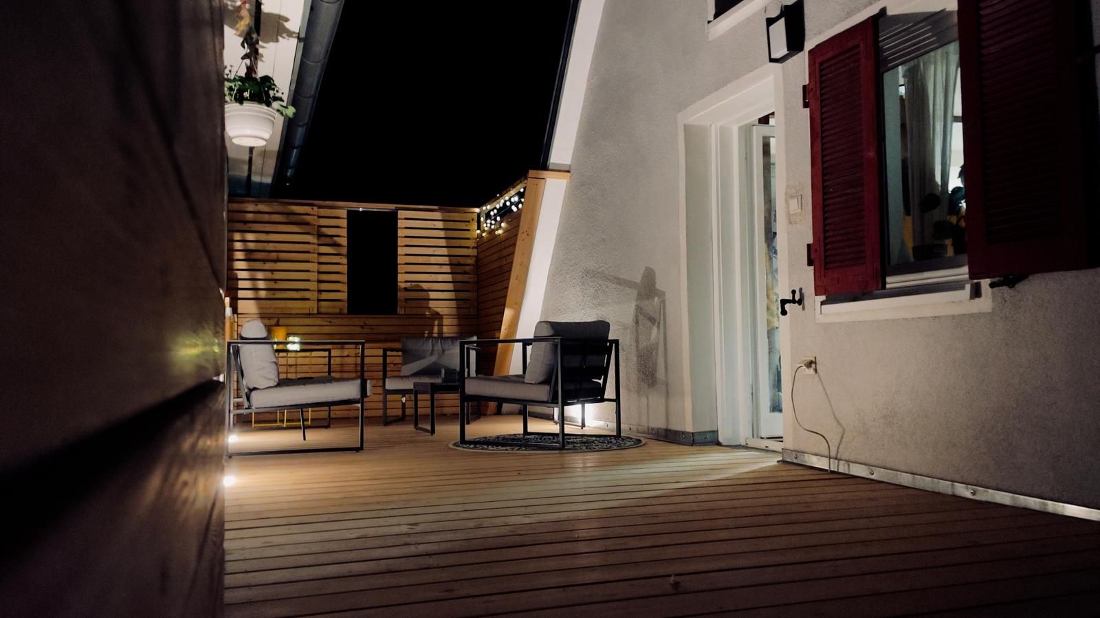 Abendliche Terrasse mit Holzboden, Gartenstühlen und offenem Fenster mit roten Fensterläden.