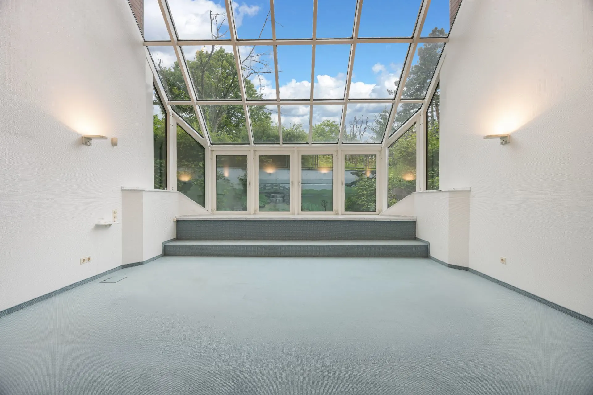 Heller Raum mit großen Glasfenstern und Glasdecke, Blick auf Bäume, Stufen führen zu Terrasse.
