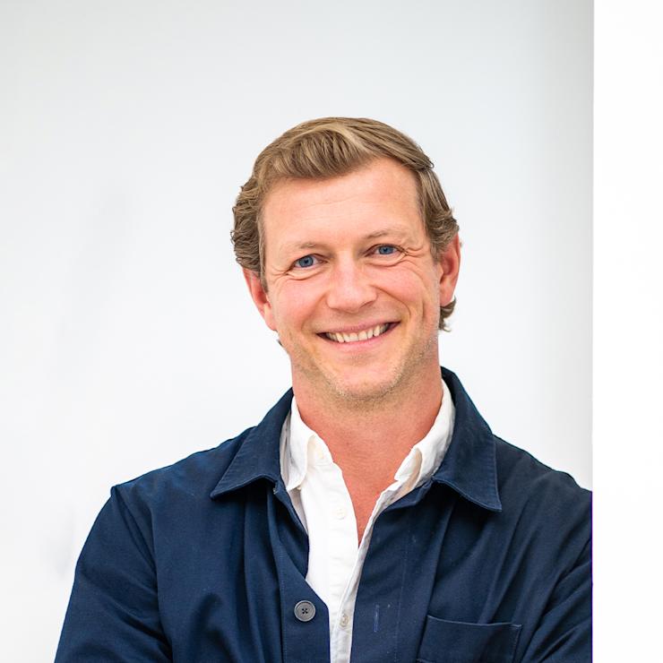 Ein Mann mit blauem Sakko und weißem Hemd steht vor einer weißen Wand und lächelt.