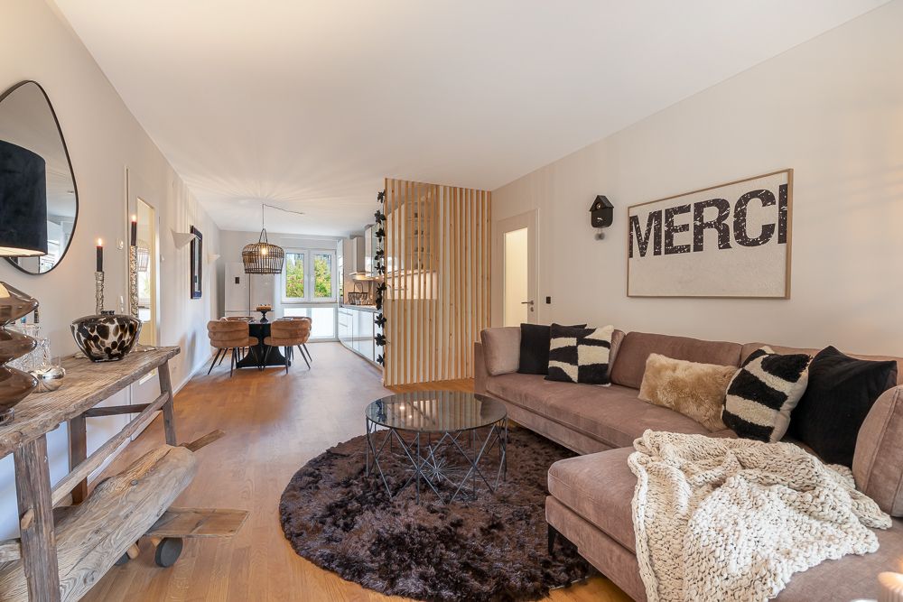 Modernes Wohnzimmer mit braunem Sofa, Glastisch und Dekor, Essbereich und Küche im Hintergrund.