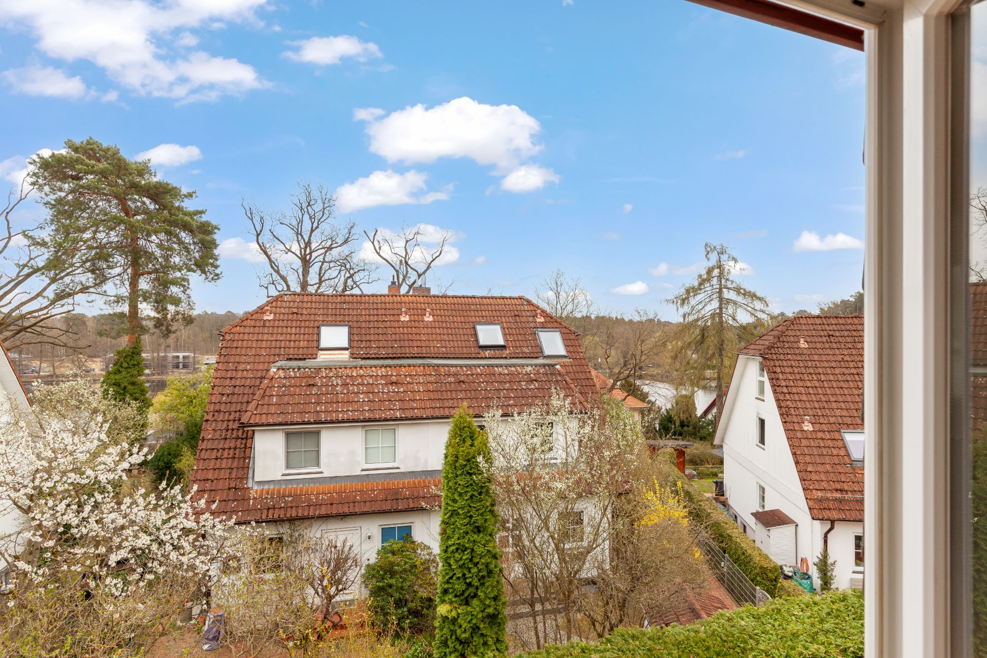 Blick aus dem Fenster auf ein rotes Wohnhaus mit Garten und Bäumen bei blauem Himmel.