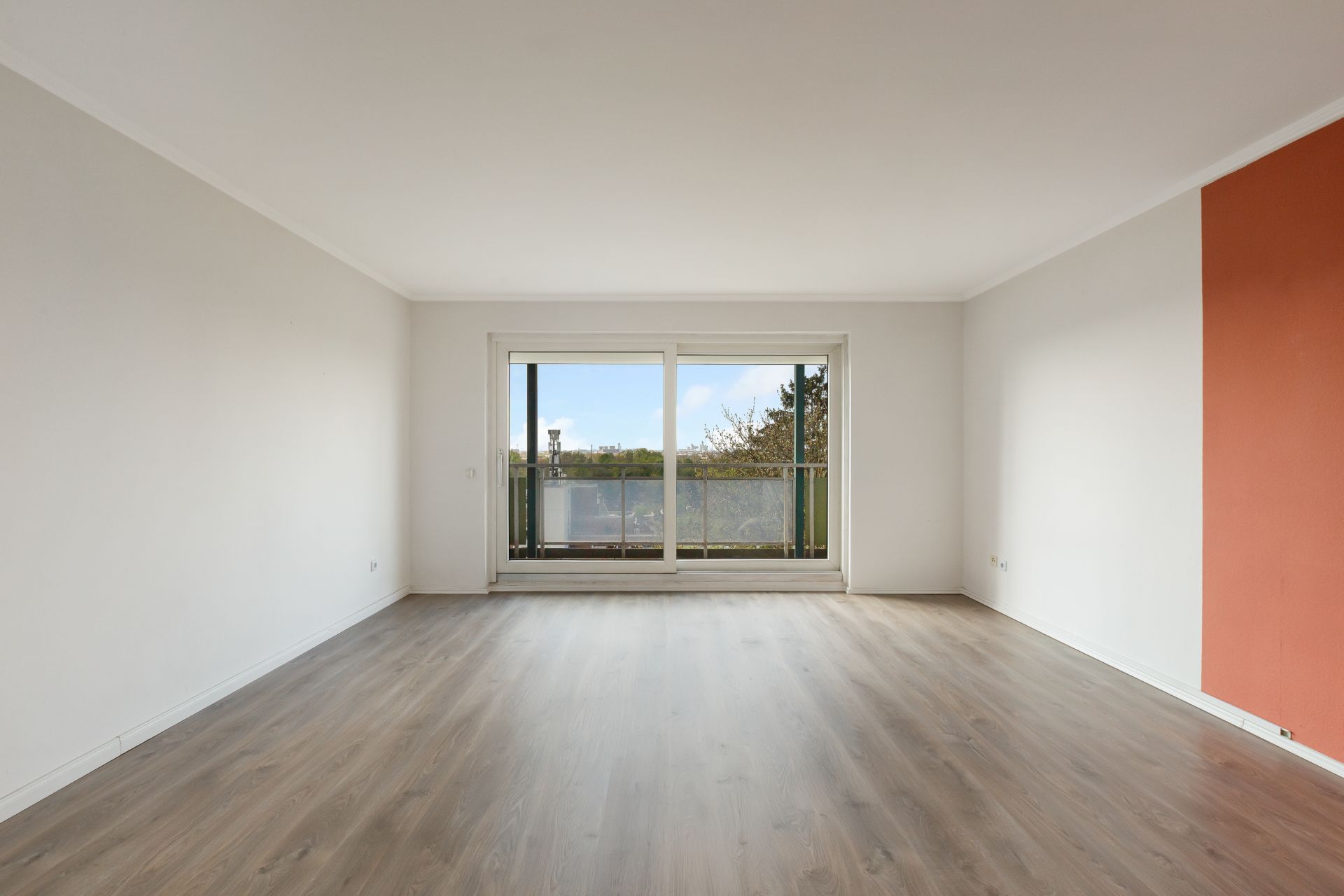 Leeres Wohnzimmer mit Holzboden, weißen Wänden und großer Fensterfront mit Zugang zum Balkon.