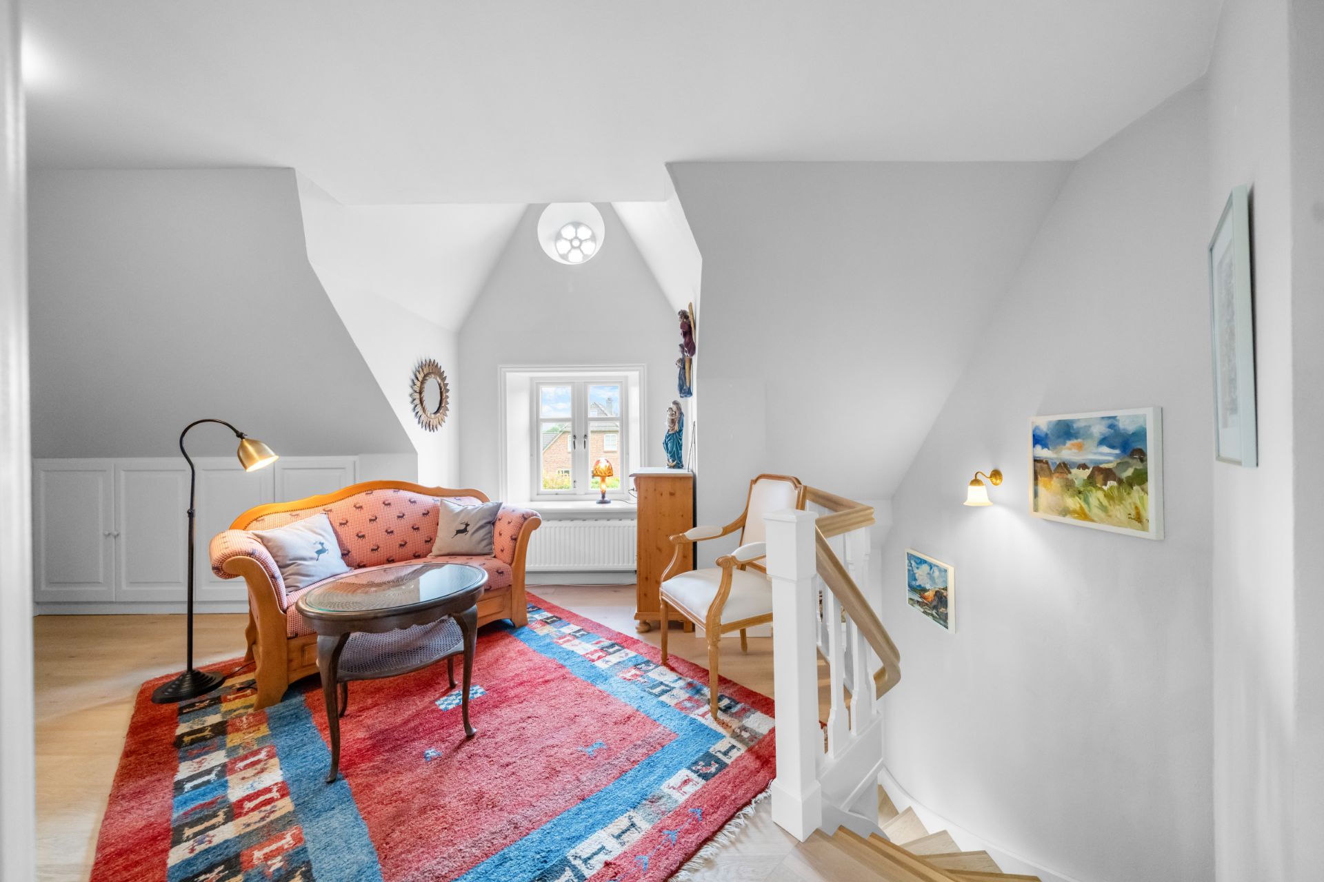 Heller Wohnbereich mit Sofa, Sessel, Tisch und farbigem Teppich neben einer Treppe.
