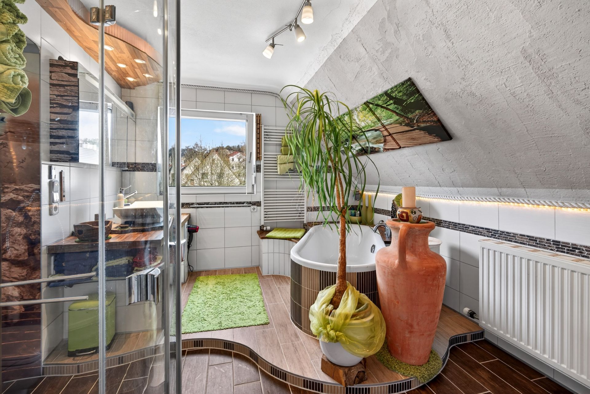 Modernes Badezimmer mit Badewanne, Dusche, Pflanze, großem Fenster und grünen Teppichen.