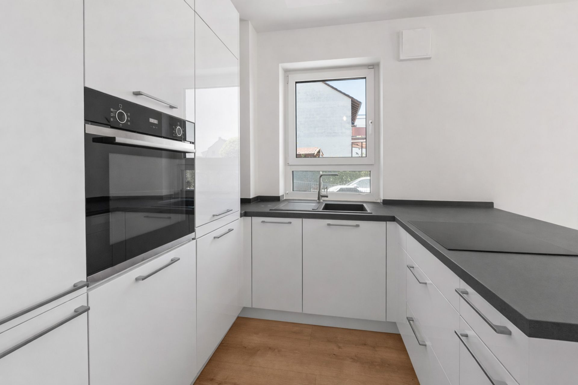 Moderne weiße Einbauküche mit Ofen, Spüle und Fenster, Holzboden sichtbar.