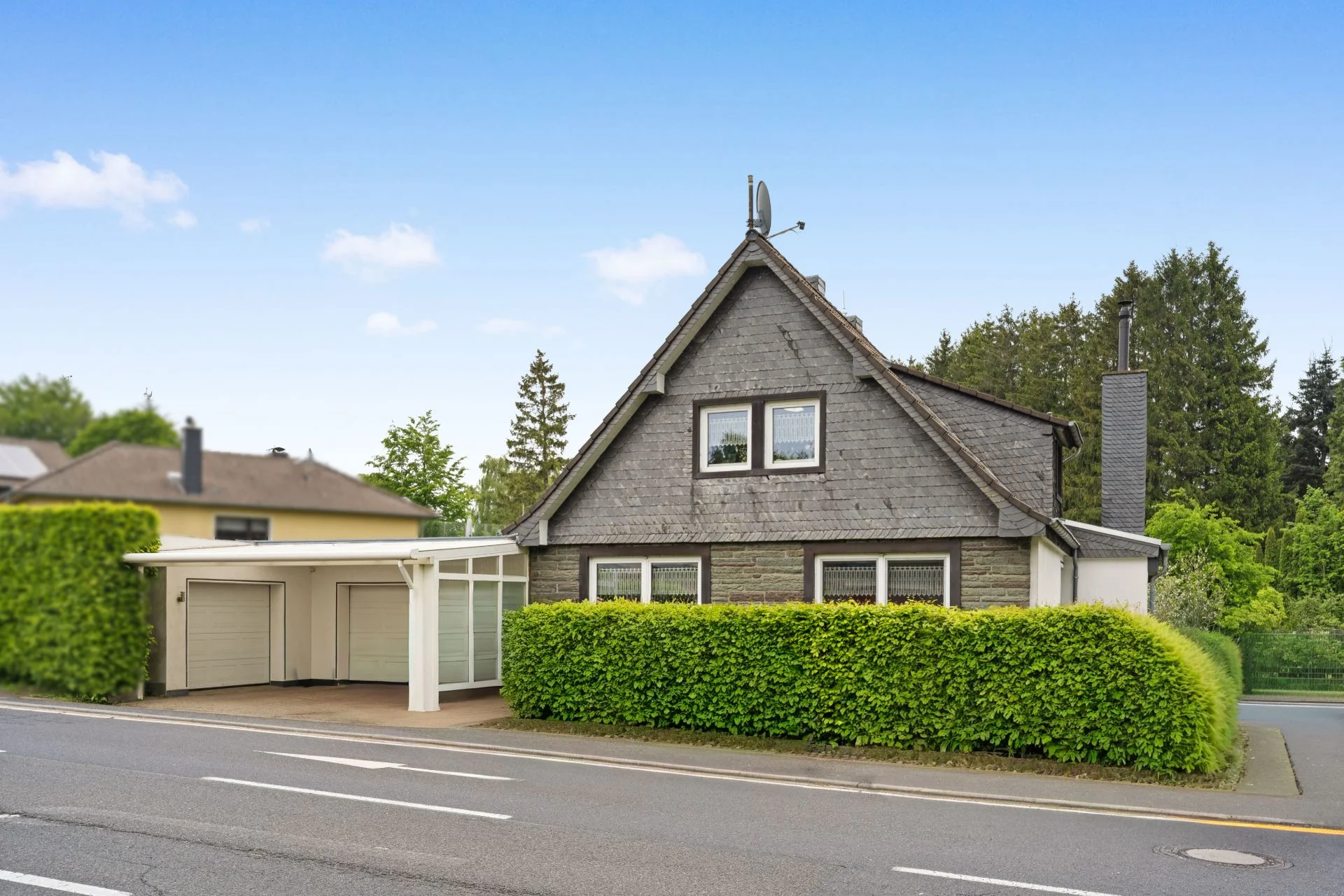 Einfamilienhaus mit spitzem Dach, Vorgartenhecke und Doppelgarage an einer Straße.