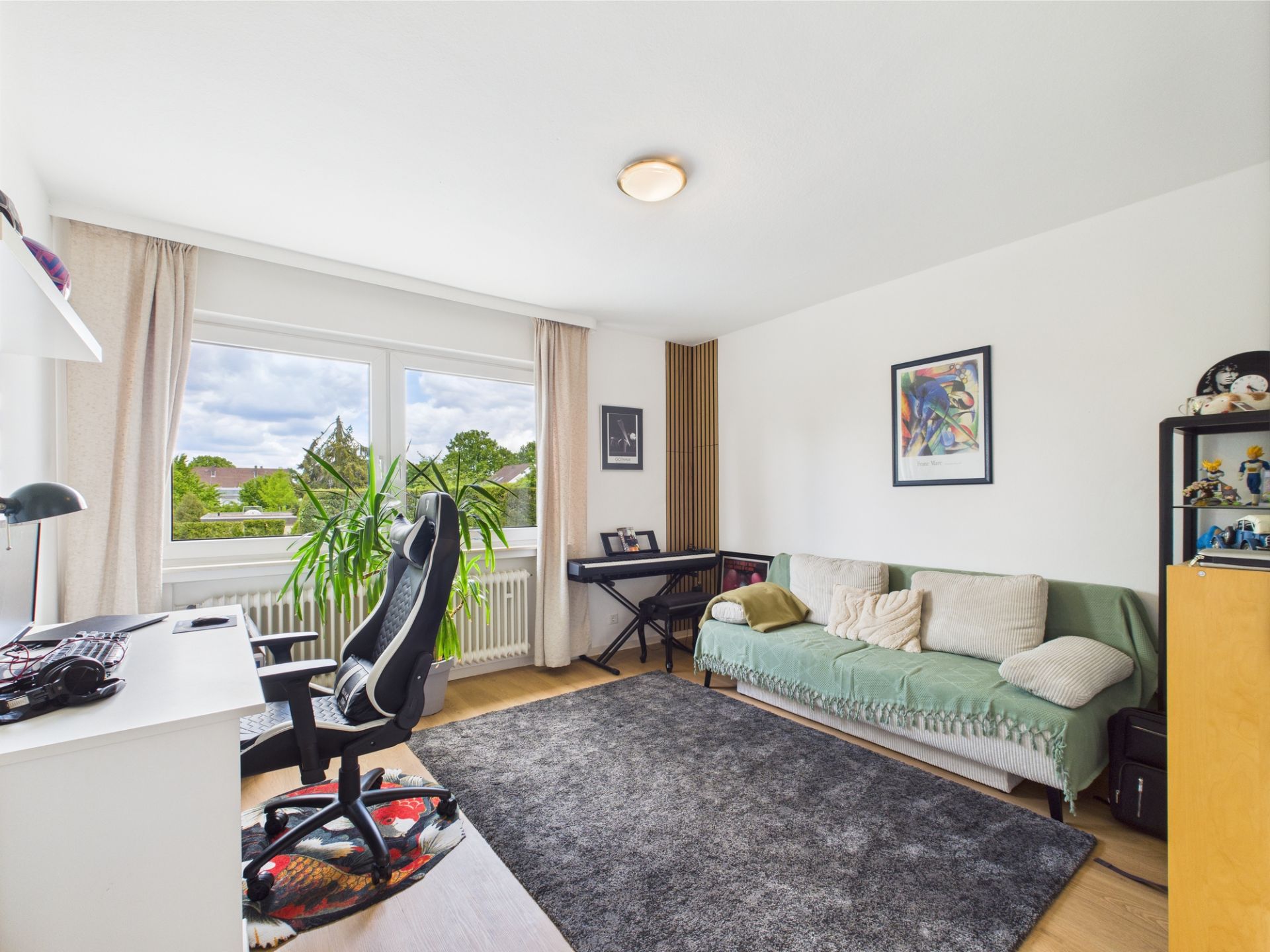 Helles Zimmer mit Schreibtisch, Bürostuhl, Sofa, Teppich und großem Fenster mit Blick ins Grüne.