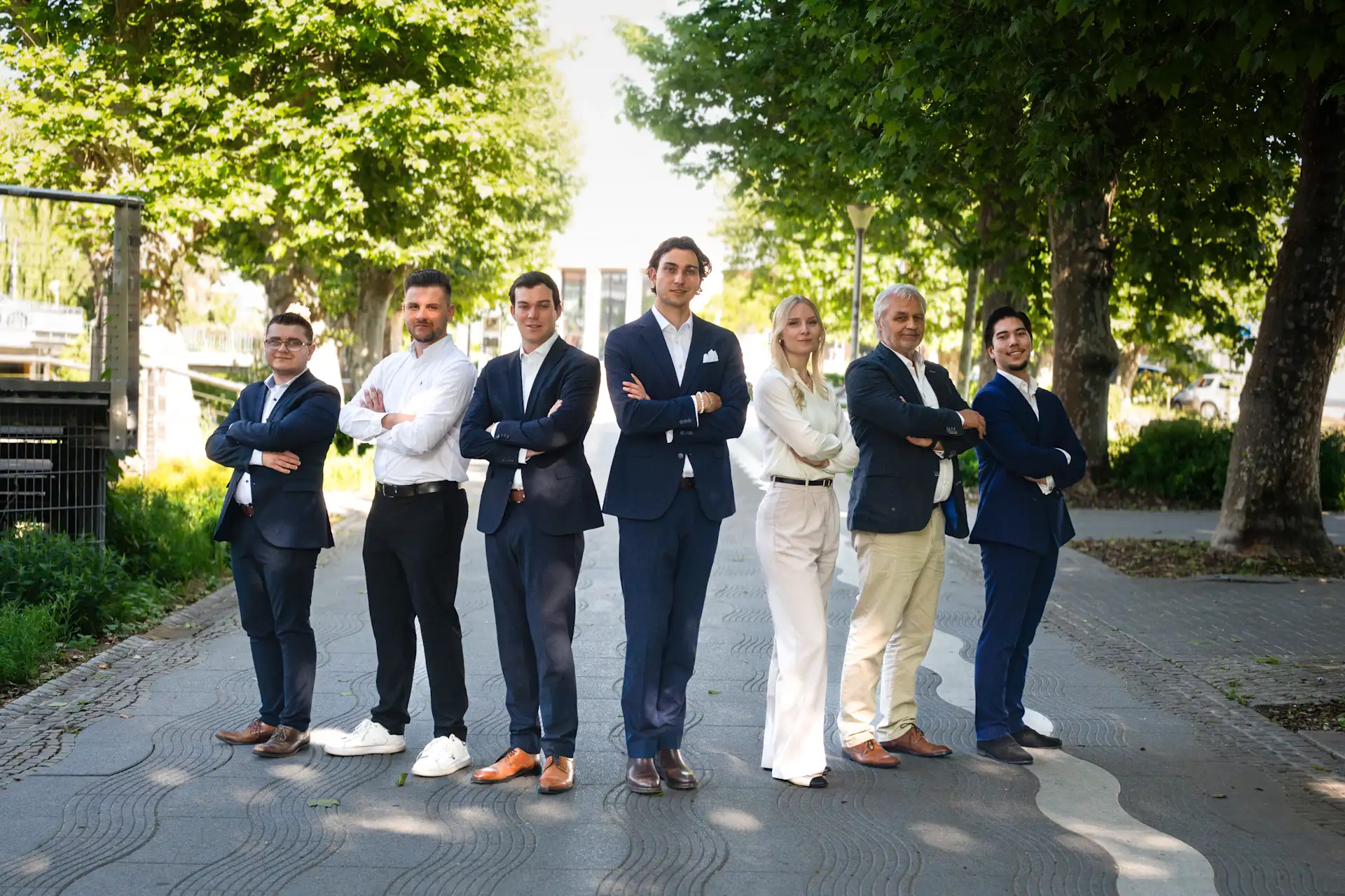 Sieben Menschen in Businesskleidung stehen draußen auf einem Gehweg mit Bäumen im Hintergrund.