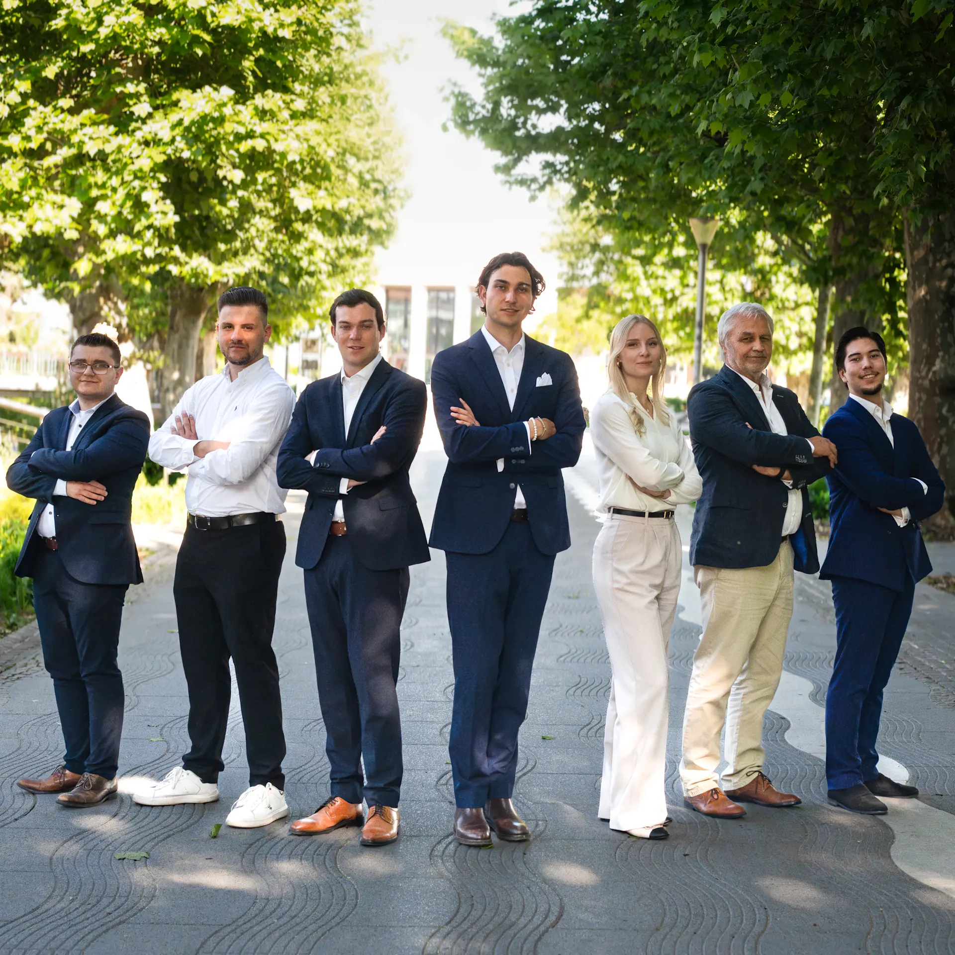 Sieben Menschen in Businesskleidung stehen draußen auf einem Gehweg mit Bäumen im Hintergrund.