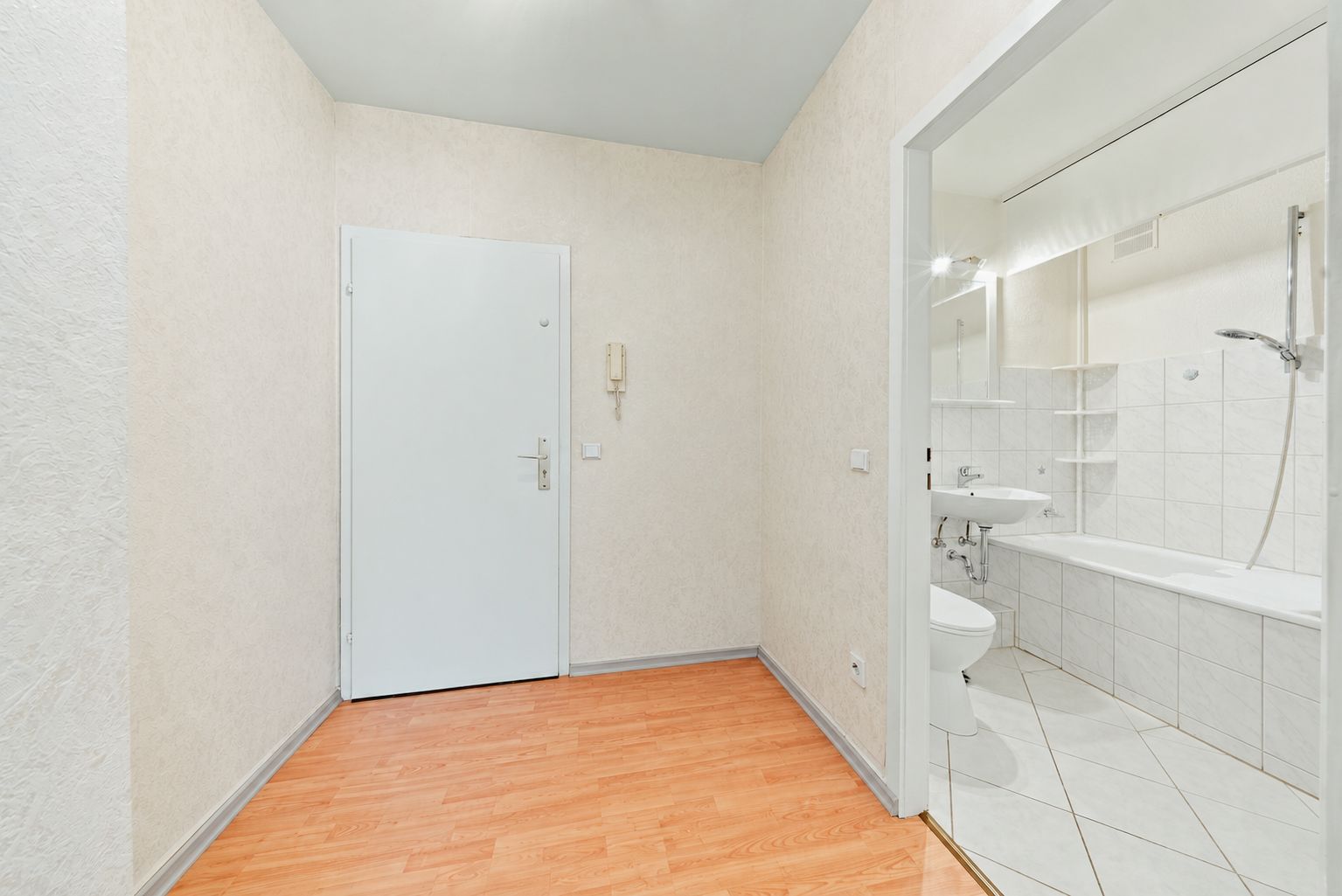 Flur mit Holzboden, weißer Tür und Blick ins Badezimmer mit Badewanne und Waschbecken.