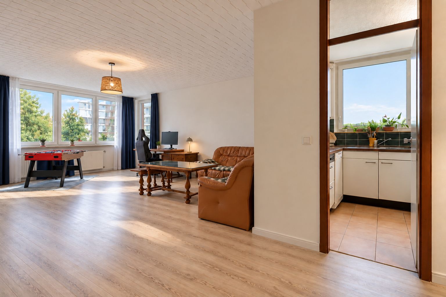 Helles Wohnzimmer mit Sofa, Schreibtisch und Blick in eine separate Küche mit großem Fenster.