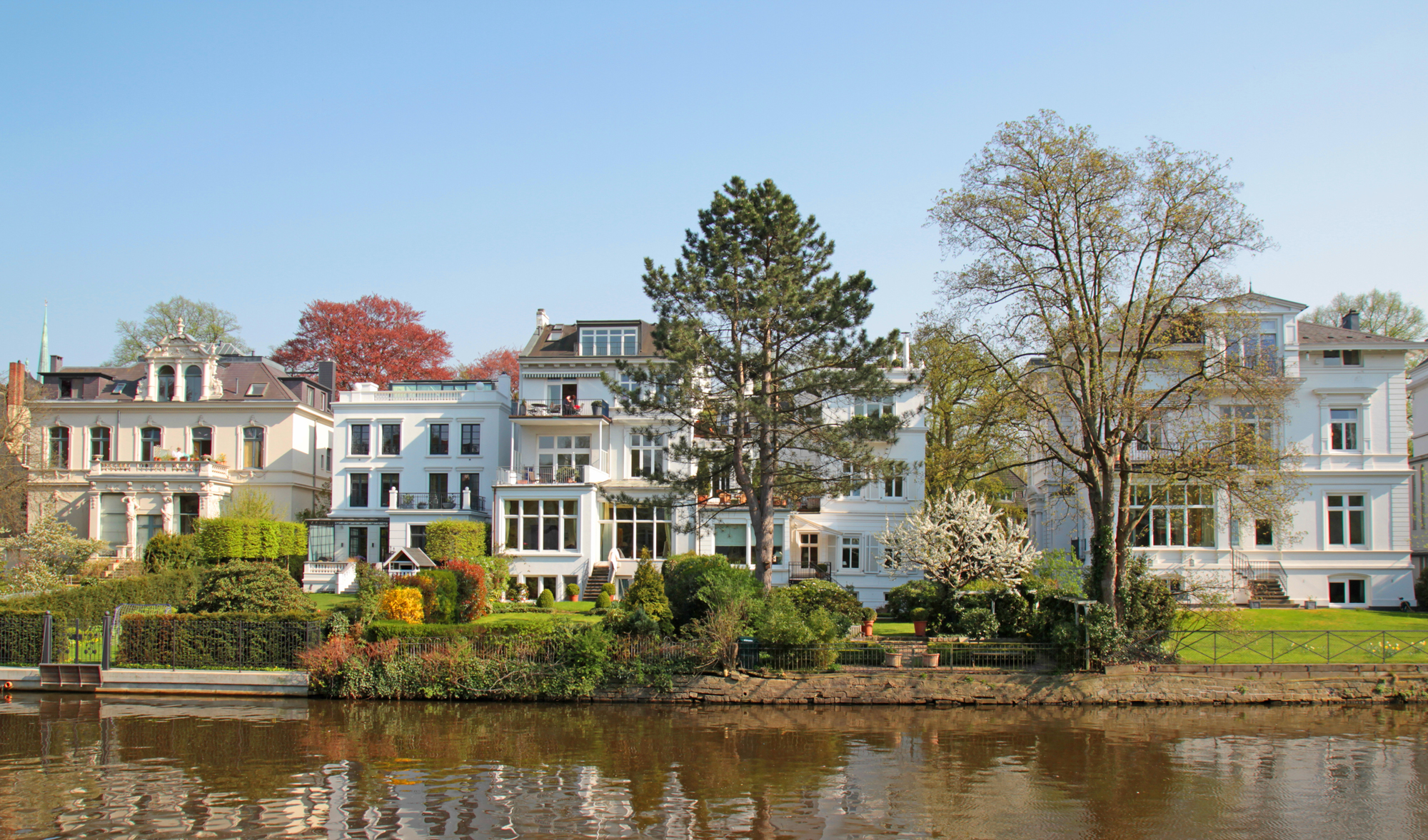 Mehrere große Wohnhäuser mit Gärten am Flussufer, Bäume im Vordergrund.