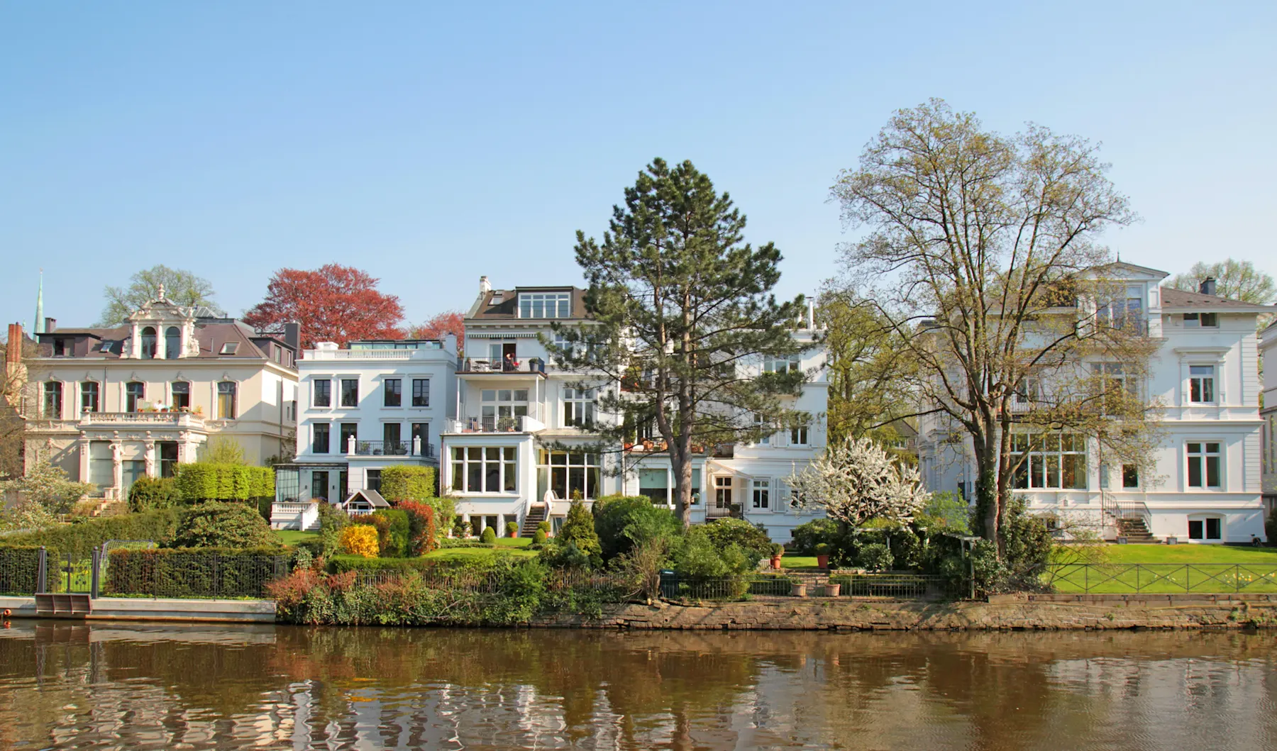 Mehrere große Wohnhäuser mit Gärten am Flussufer, Bäume im Vordergrund.