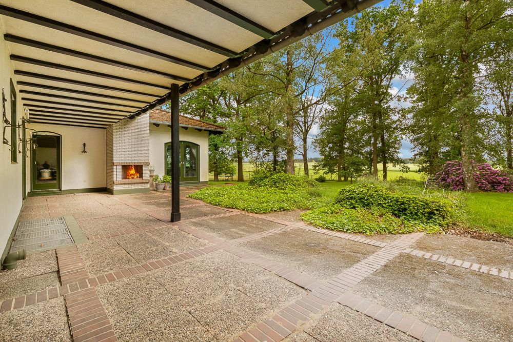Überdachte Terrasse mit offenem Kamin, Blick auf Garten mit Bäumen und Rasen.