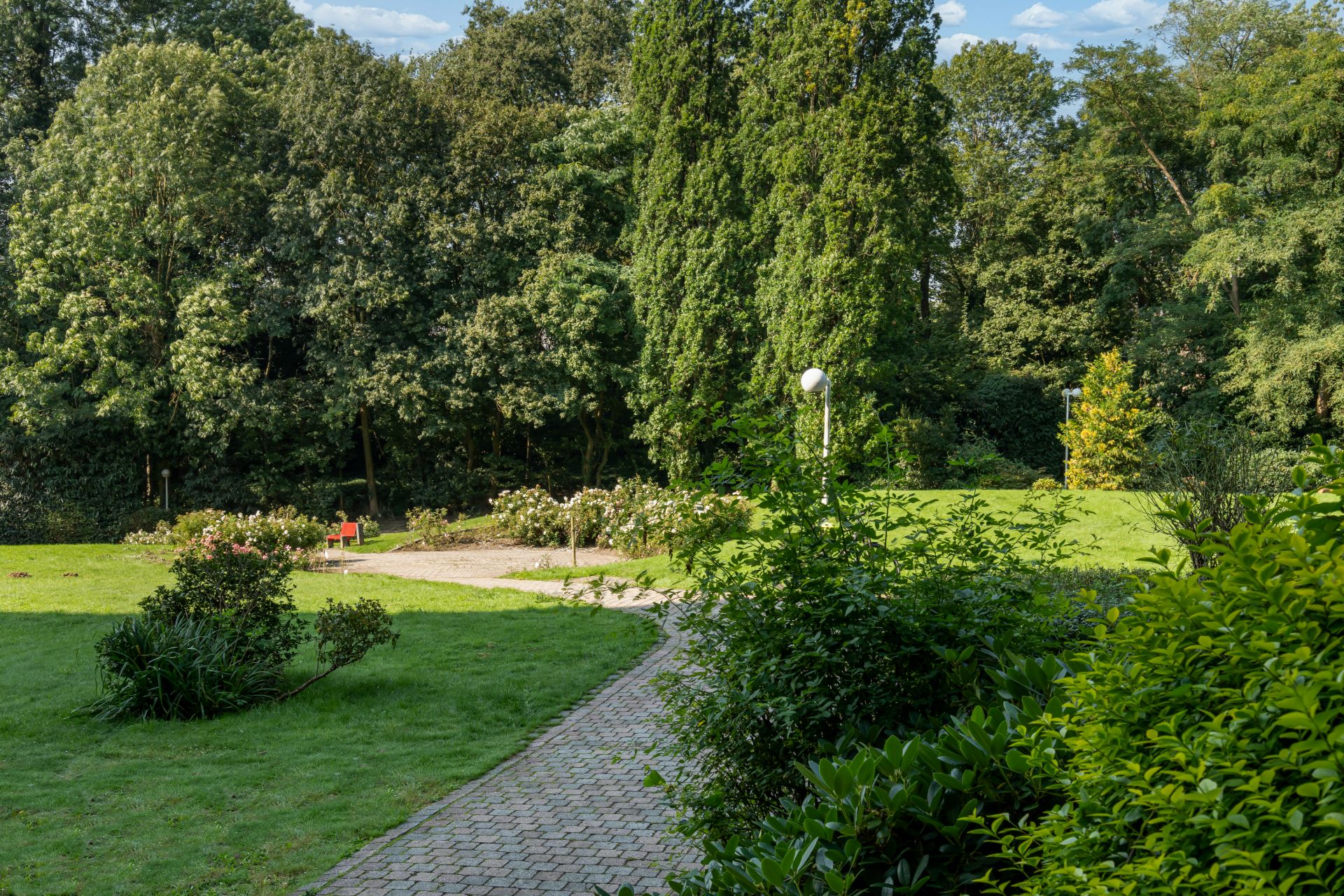 Gepflasterter Weg durch einen grünen Garten mit Büschen, Rasen und Bäumen im Hintergrund.