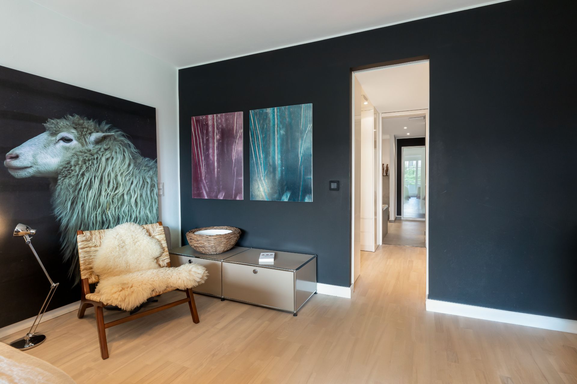 Modernes Wohnzimmer mit schwarzer Wand, Stuhl mit Fell, großem Schafbild und bunten Wandbildern.