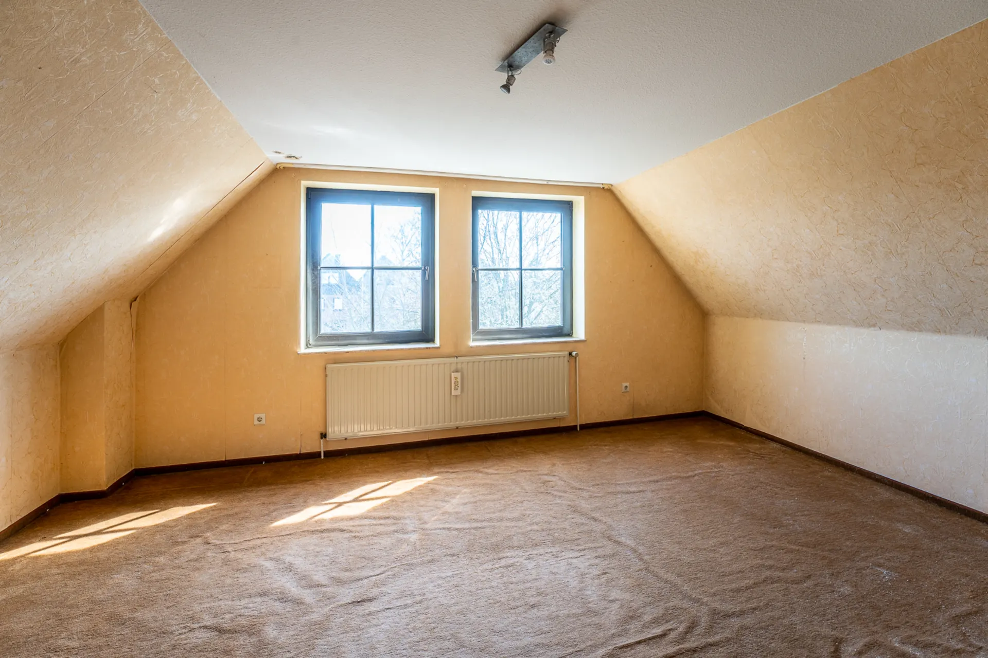 Leerstehender Dachzimmerraum mit zwei Fenstern, Teppichboden und Heizkörper unter den Fenstern.