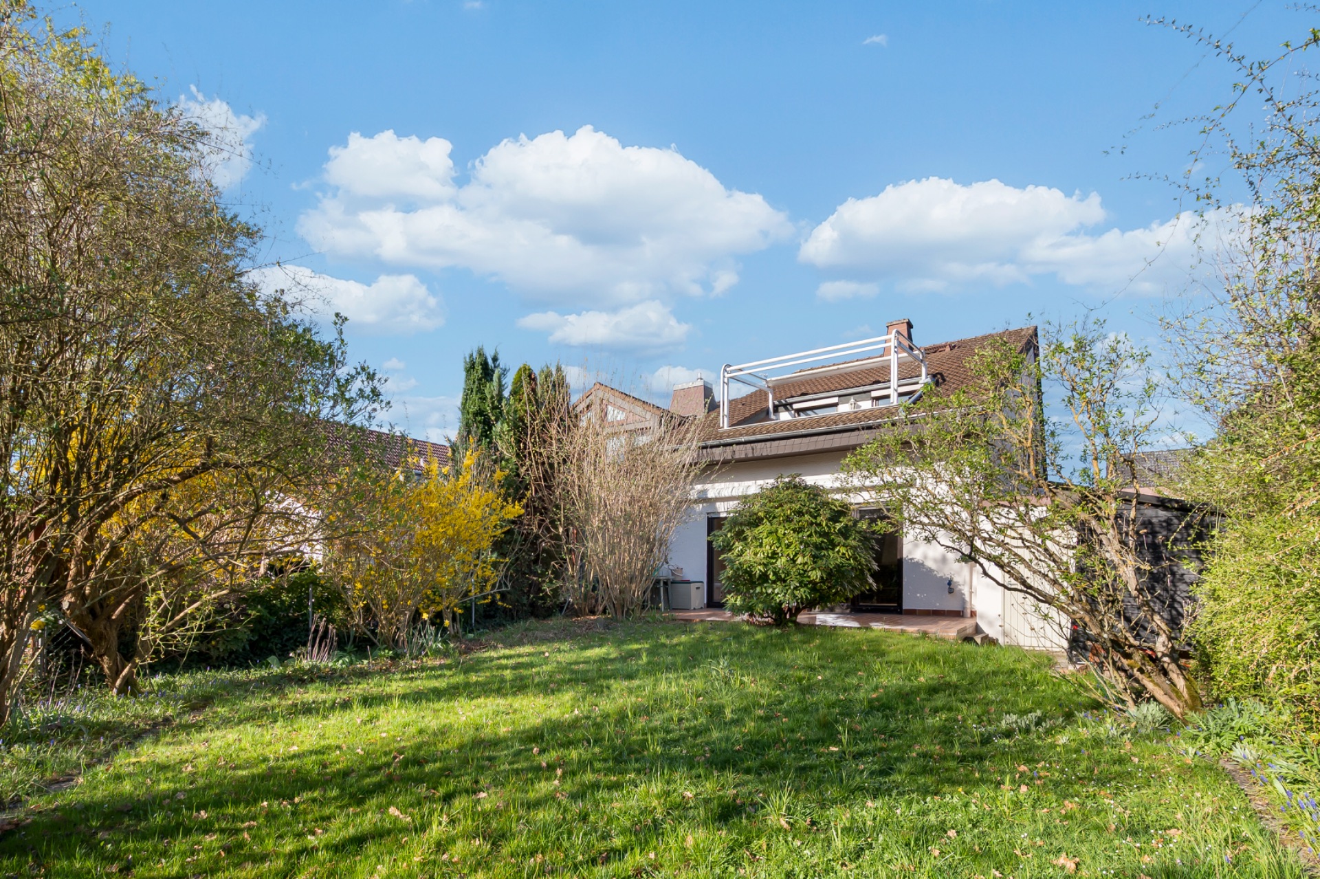 Haus mit Terrasse und großem Garten, umgeben von Bäumen und Sträuchern, unter blauem Himmel.