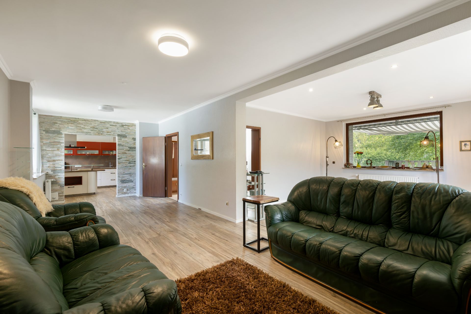 Wohnzimmer mit zwei grünen Ledersofas, Teppich und Blick in eine offene Küche mit Steinwand.