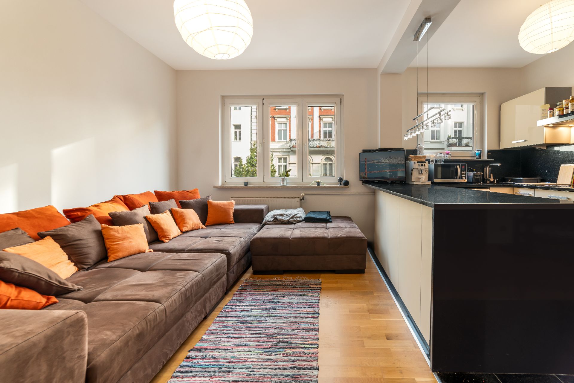 Wohnzimmer mit braunem Sofa, bunten Kissen und offener Küche, große Fenster im Hintergrund.
