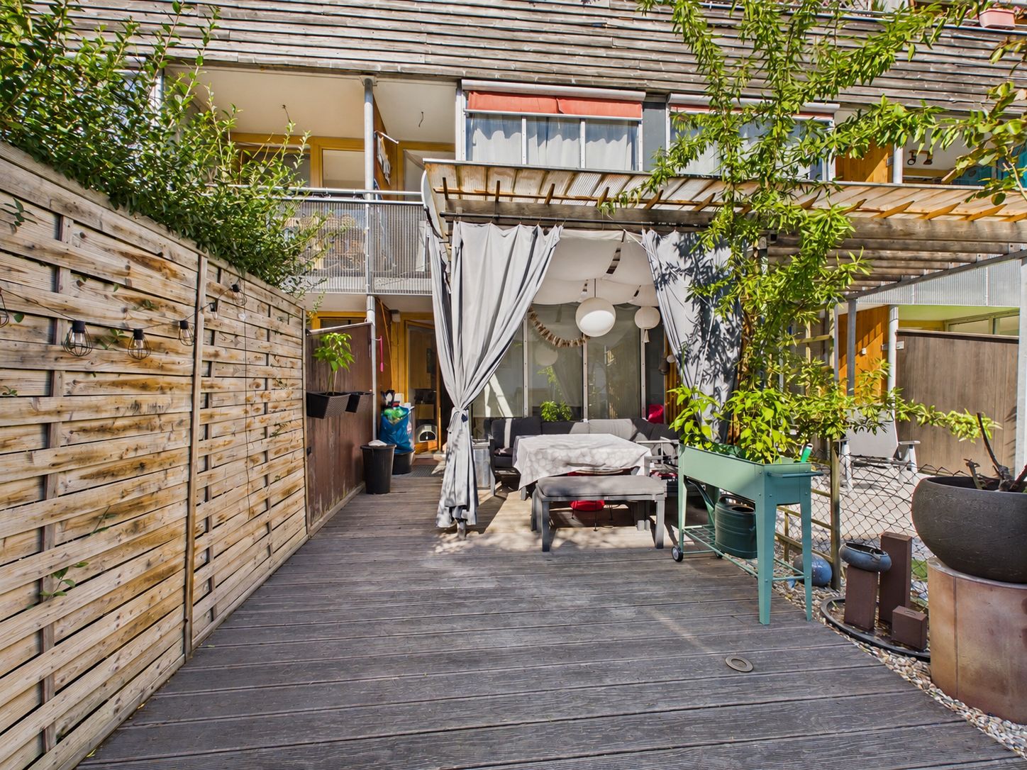 Holzterrasse mit Pergola, weißen Vorhängen, Sitzmöbeln und Pflanzen in einem Innenhof.