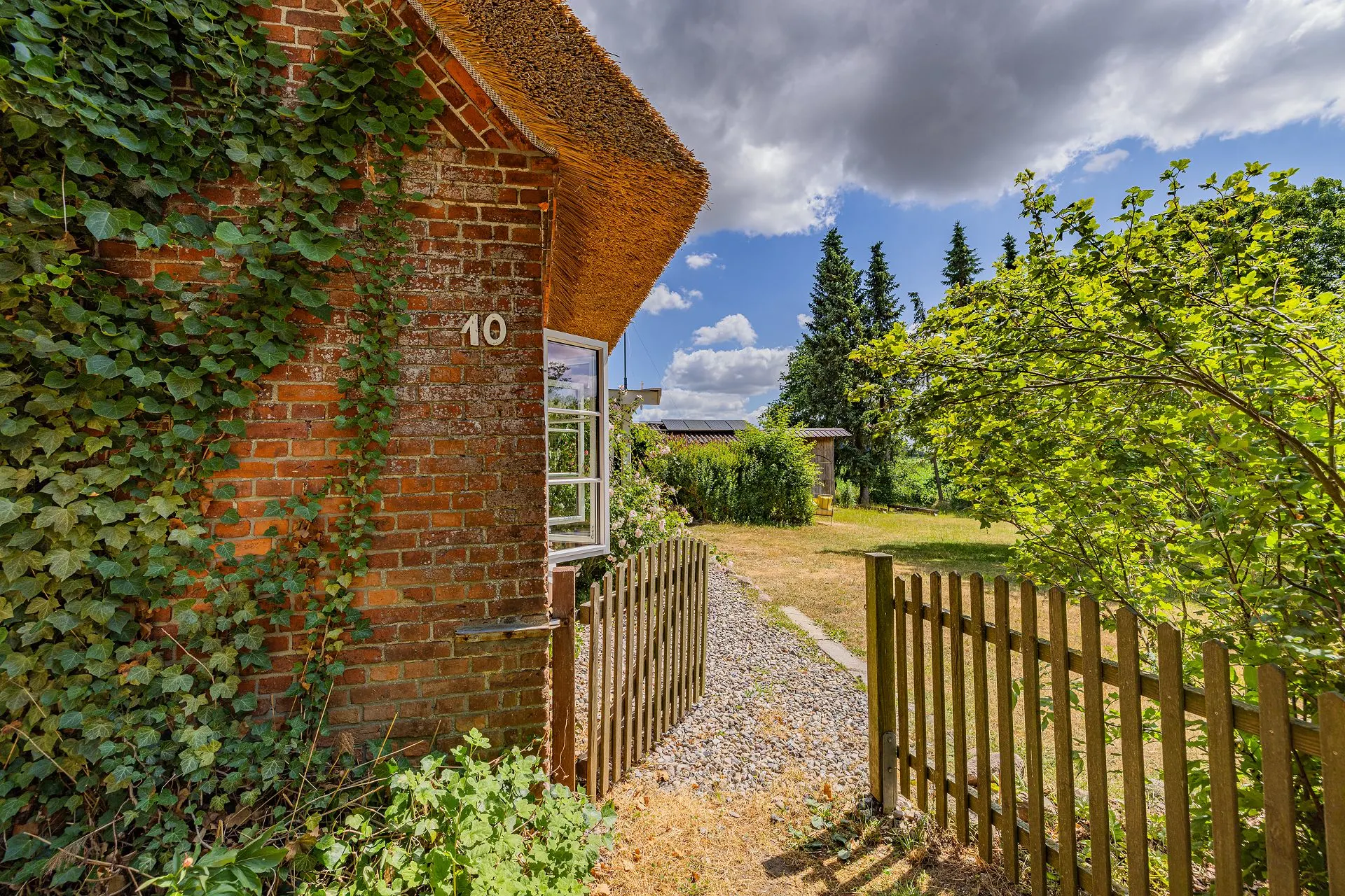 Backsteinhaus mit Reetdach, offenem Fenster, Zaun und grünem Garten im Sonnenschein.