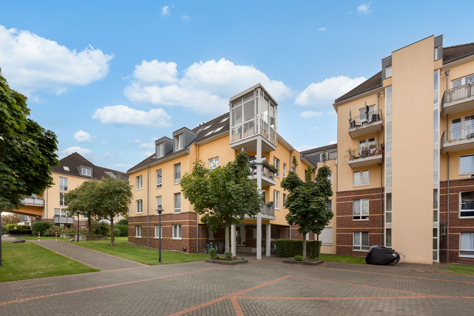 Mehrstöckiges Wohngebäude mit gelber Fassade, umliegenden Bäumen und gepflastertem Hof bei Tageslicht.
