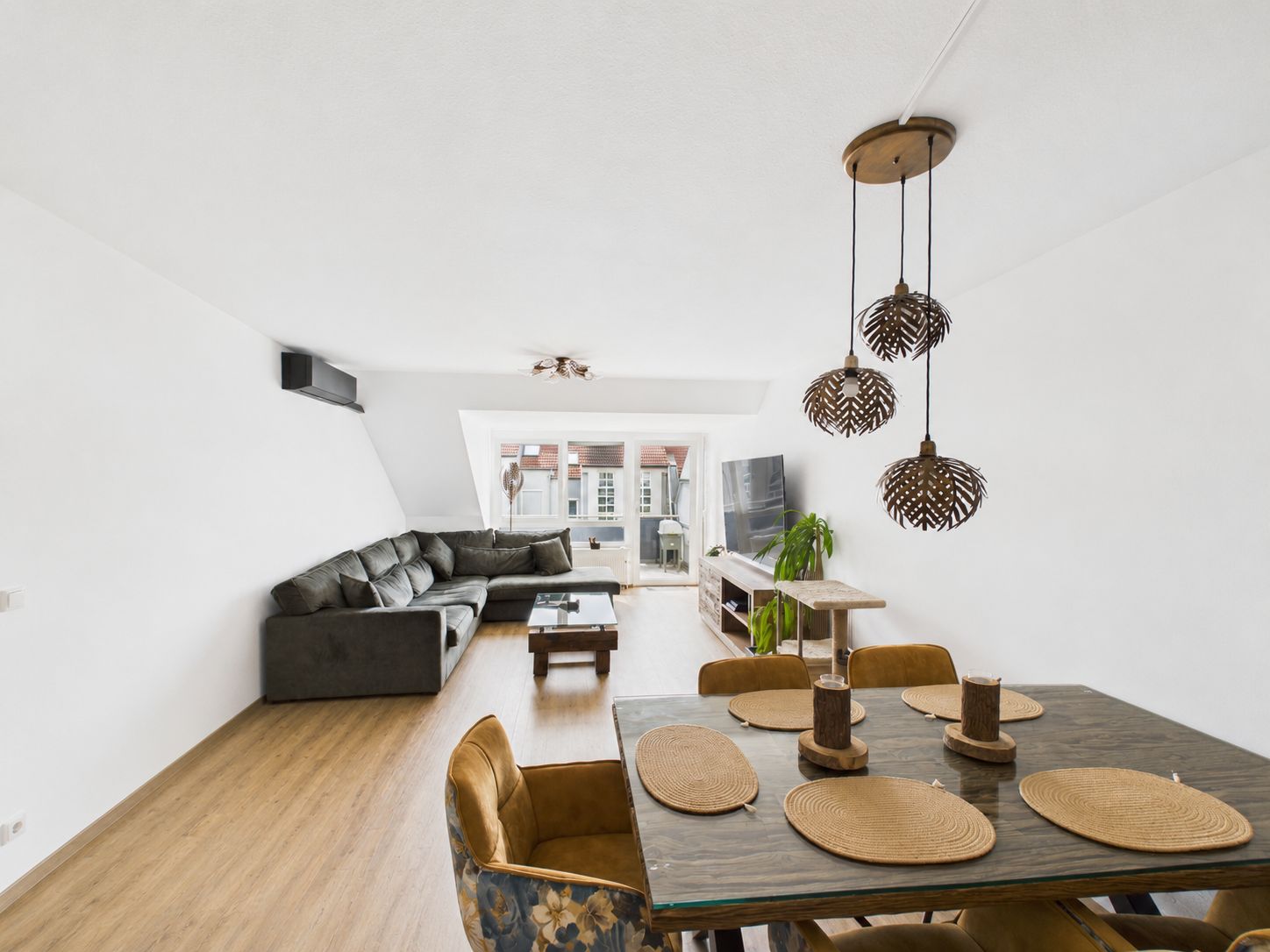 Wohnzimmer mit Ecksofa, Esstisch, Hängelampen und Fernseher, heller Holzboden.