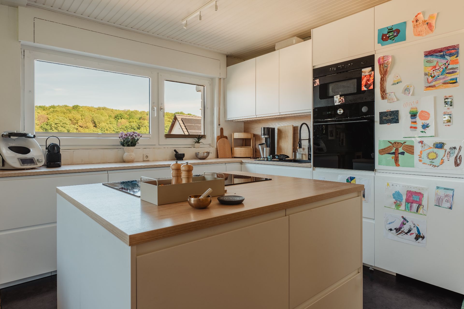 Moderne Küche mit Kochinsel, großen Fenstern und Kühlschrank mit Kinderzeichnungen.