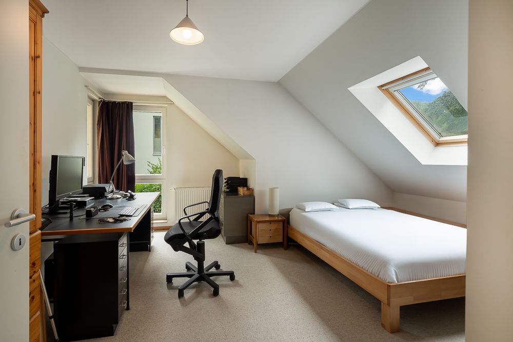 Helles Zimmer mit Schreibtisch, Bürostuhl, Bett und Dachfenster.