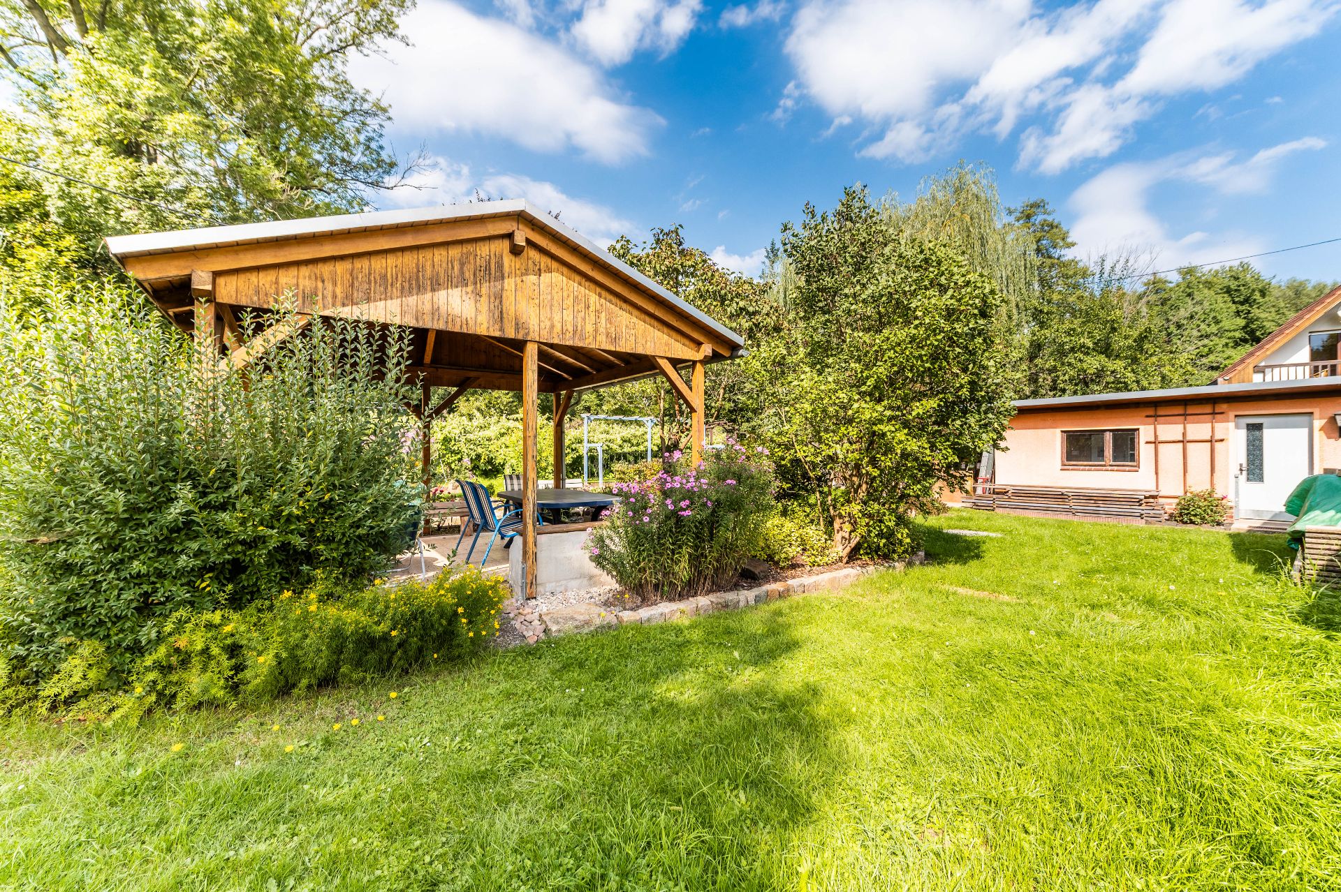 Garten mit überdachtem Holzpavillon, Sitzgruppe, viel Grün und Haus im Hintergrund.