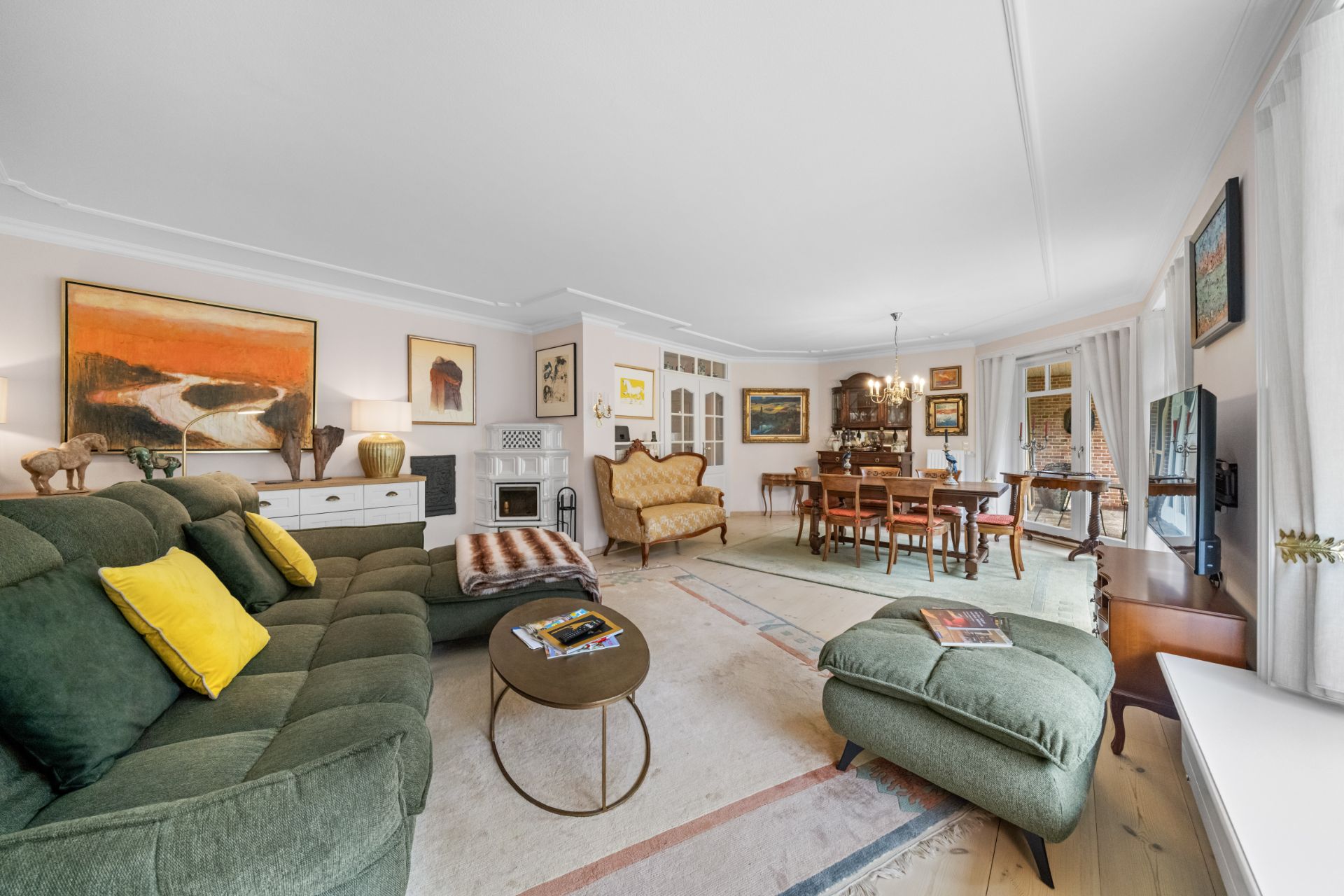 Großes Wohnzimmer mit grüner Couch, Esstisch, Gemälden und vielen Fenstern.