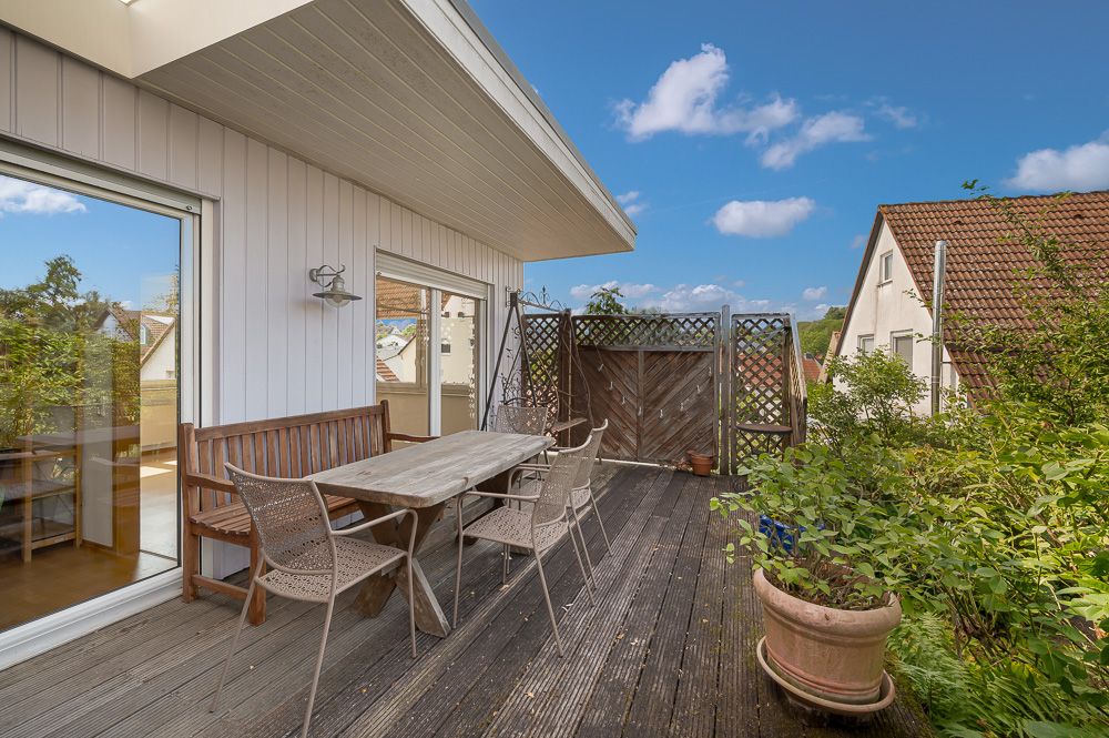 Holzterrasse mit Tisch, Stühlen, Bank und Pflanzen vor einem Wohnhaus unter blauem Himmel.