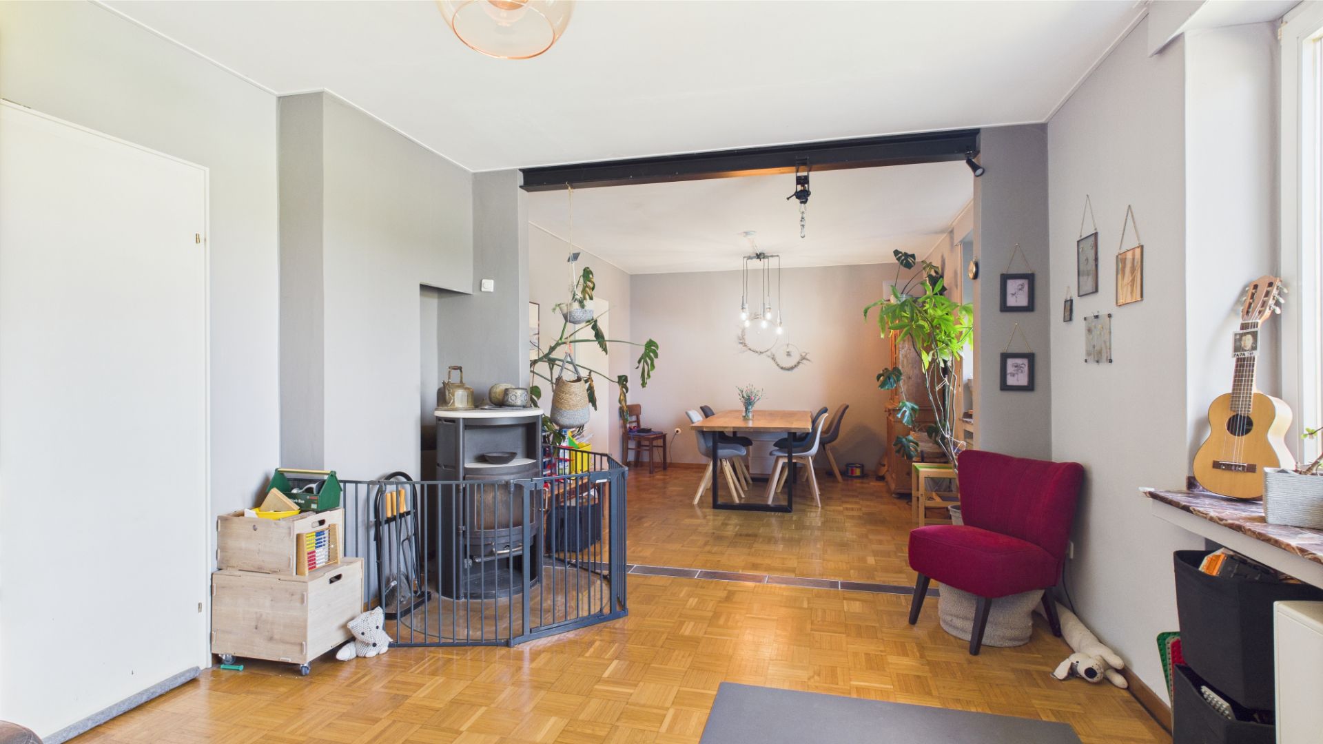 Wohnzimmer mit Holzofen, Kinderschutzgitter, Esstisch, Pflanzen und Fenster mit Gitarre auf der Fensterbank.